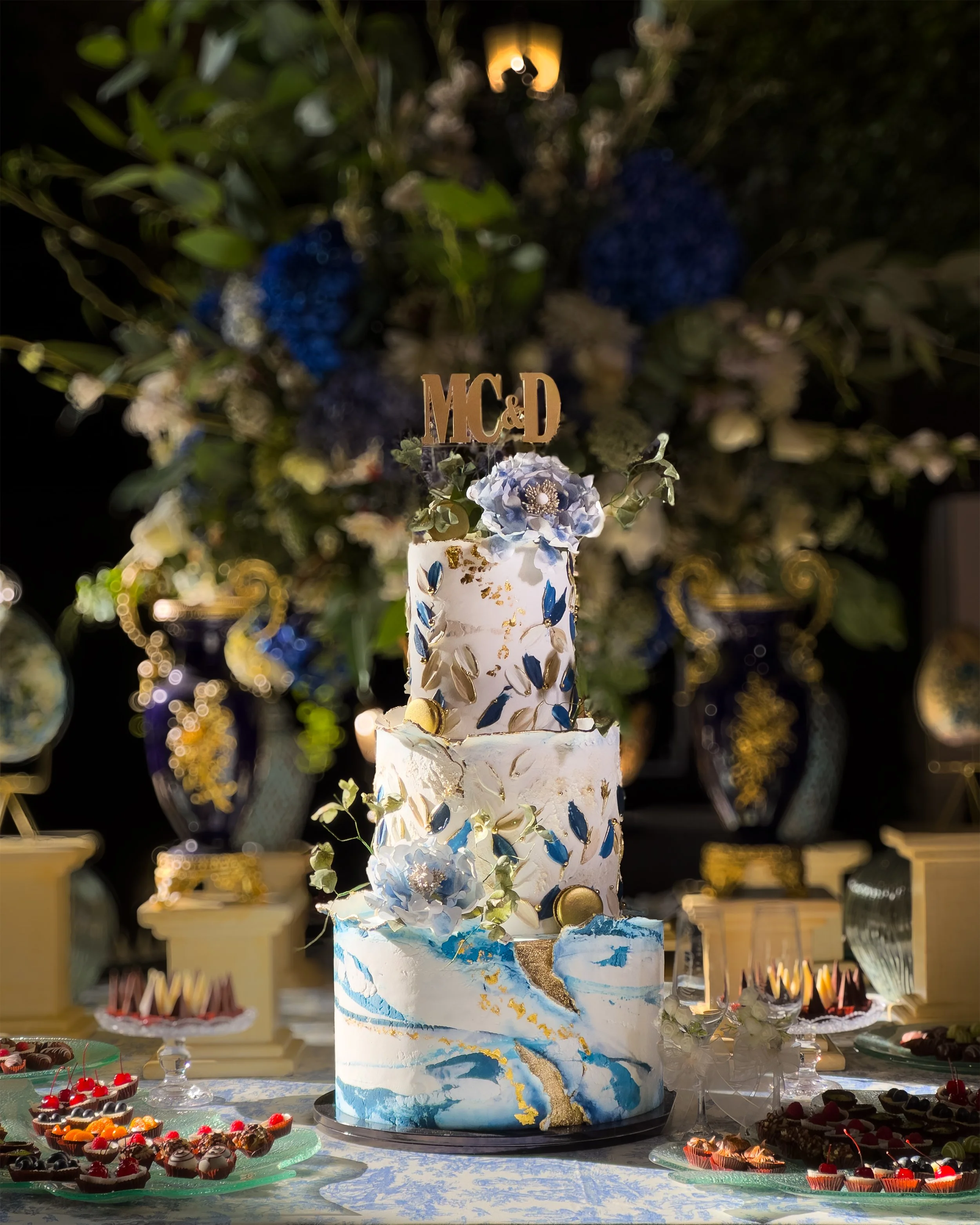 A tall, elegant four-tiered wedding cake decorated with blue, white, and gold accents, topped with a flower and a gold 'M&D' cake topper, set on a decorated table with assorted desserts, with a floral and greenery background.