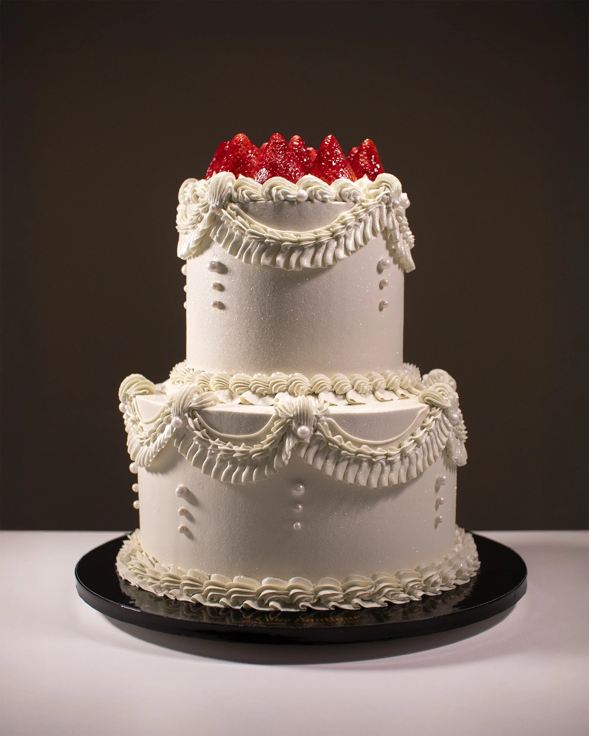 A three-tiered white wedding cake decorated with white frosting swags, pearl-like accents, and topped with fresh strawberries.