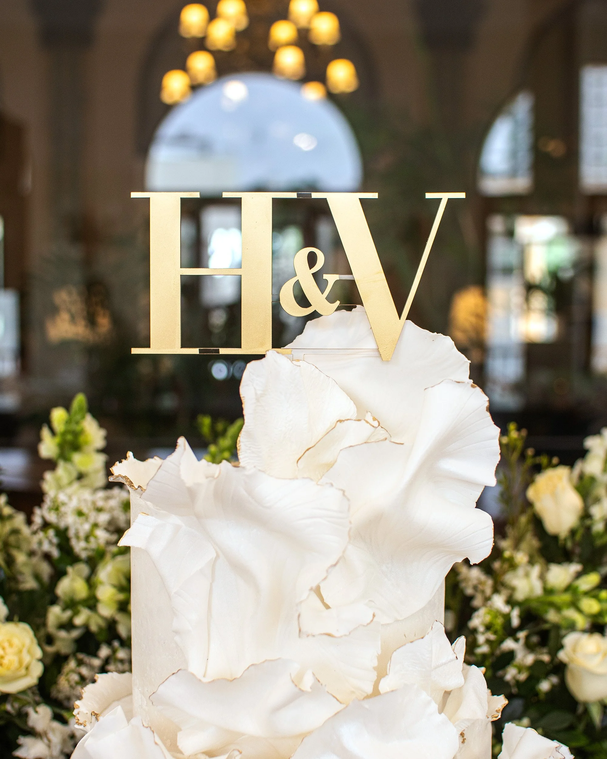 A wedding cake decorated with large white flowers and a topper with the initials 'H & V'.