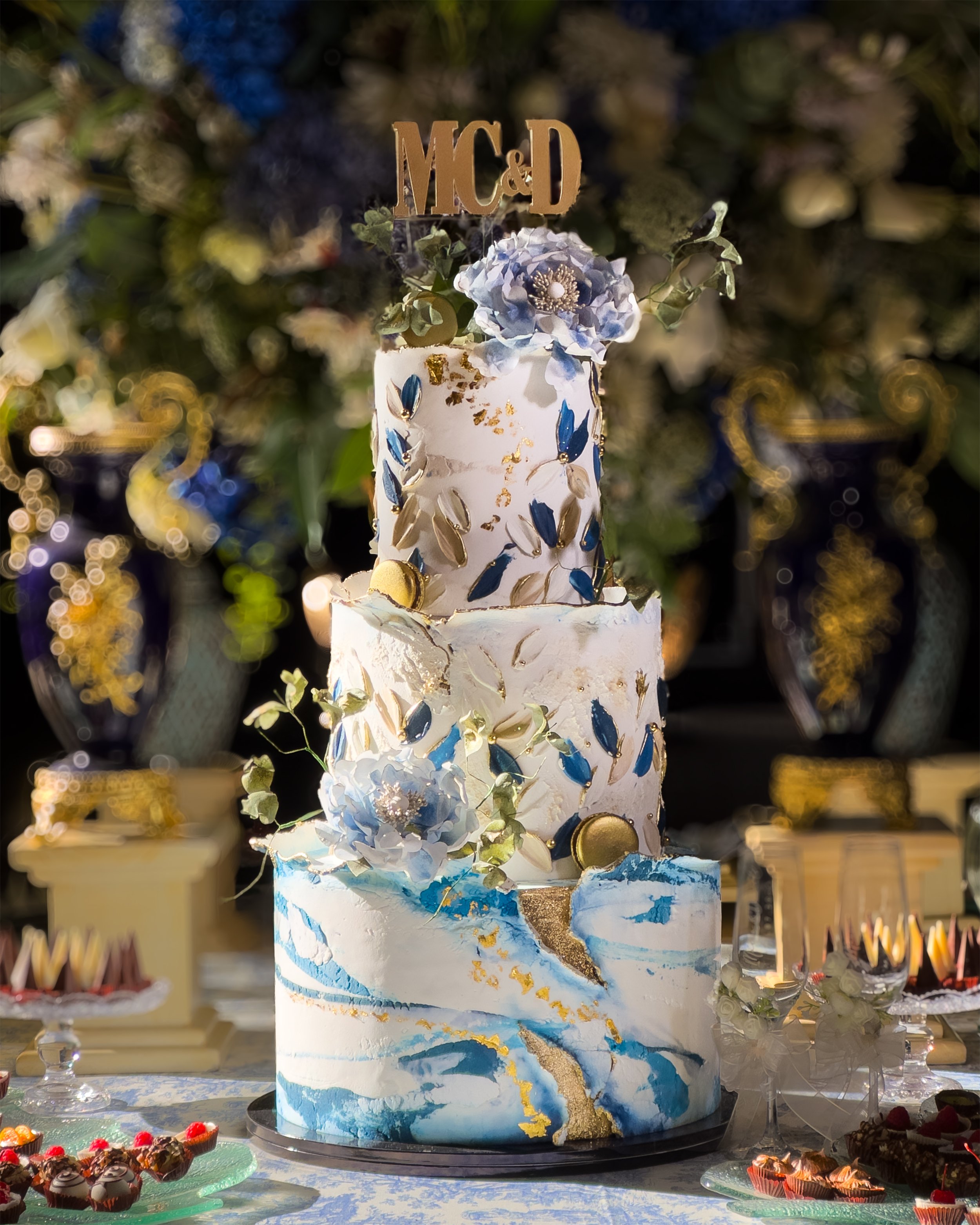A three-tiered wedding cake decorated with blue, white, and gold accents, topped with a blue flower and a gold cake topper with the initials 'MC & D', surrounded by desserts on a decorated table.