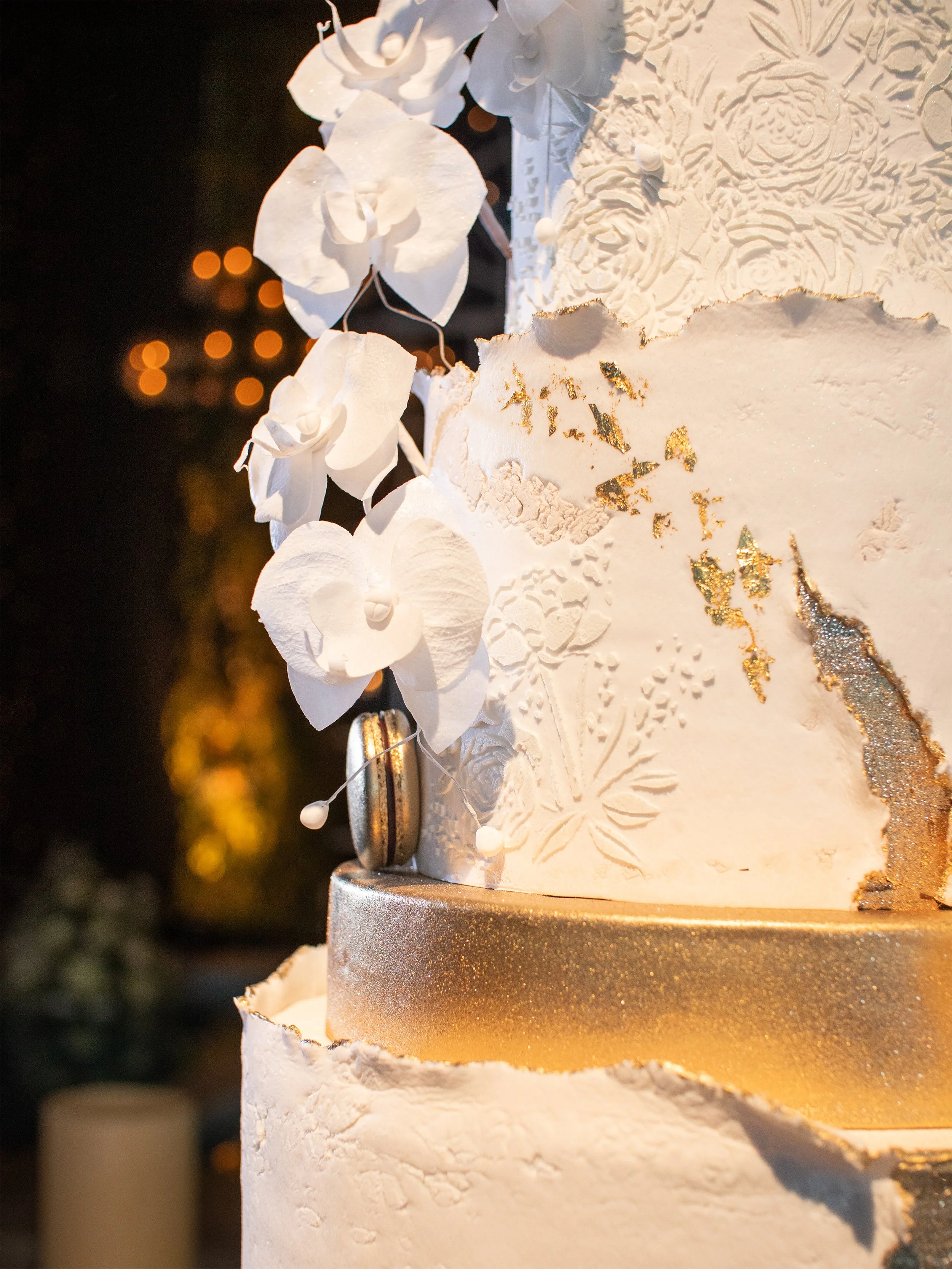 Close-up of a white wedding cake decorated with white flowers, gold glitter accents, and intricate icing patterns, with blurred background lights.