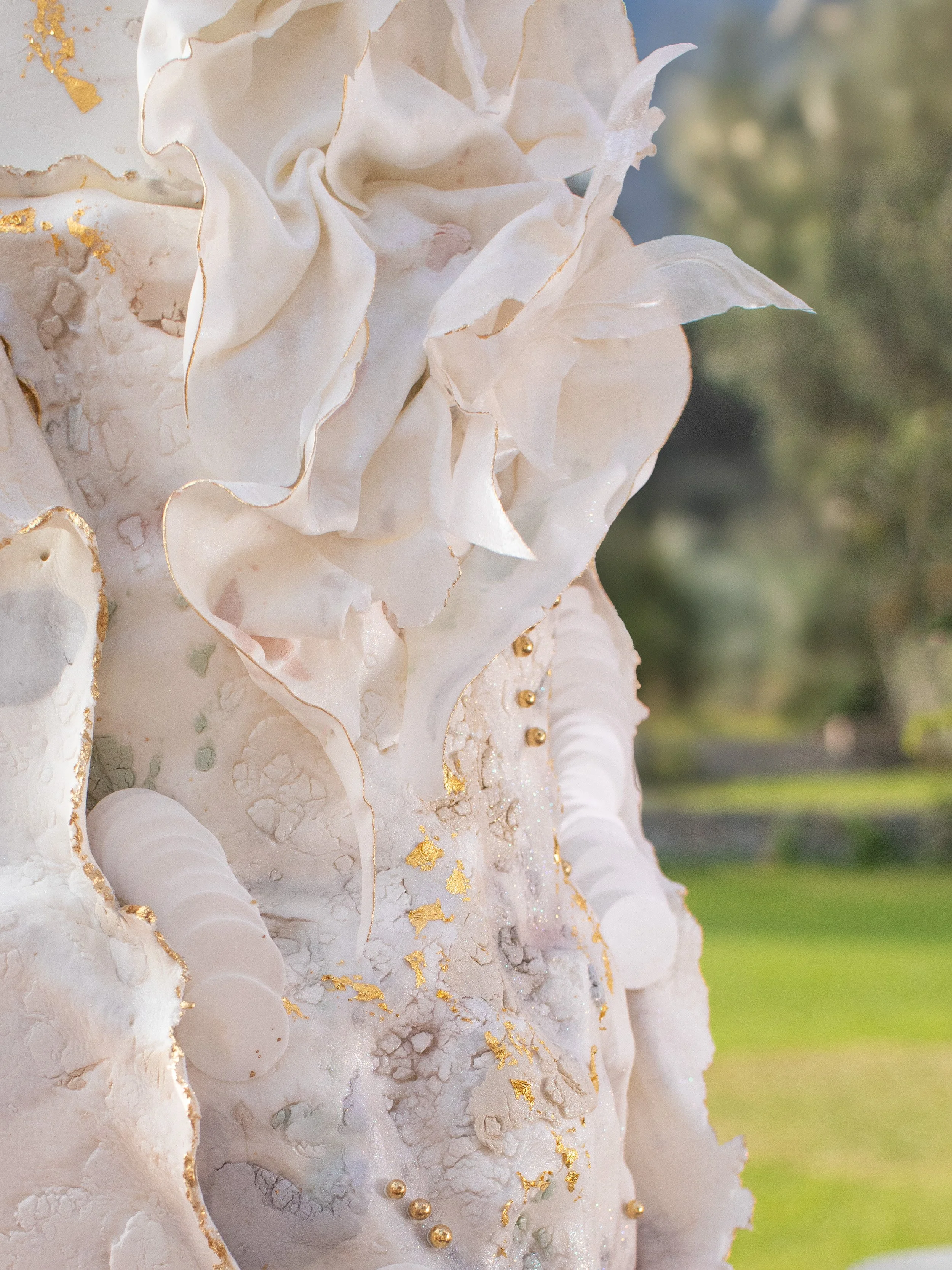 Close-up of an intricate white sculpture with gold accents, featuring textured and ruffled details, set against a blurred outdoor background.