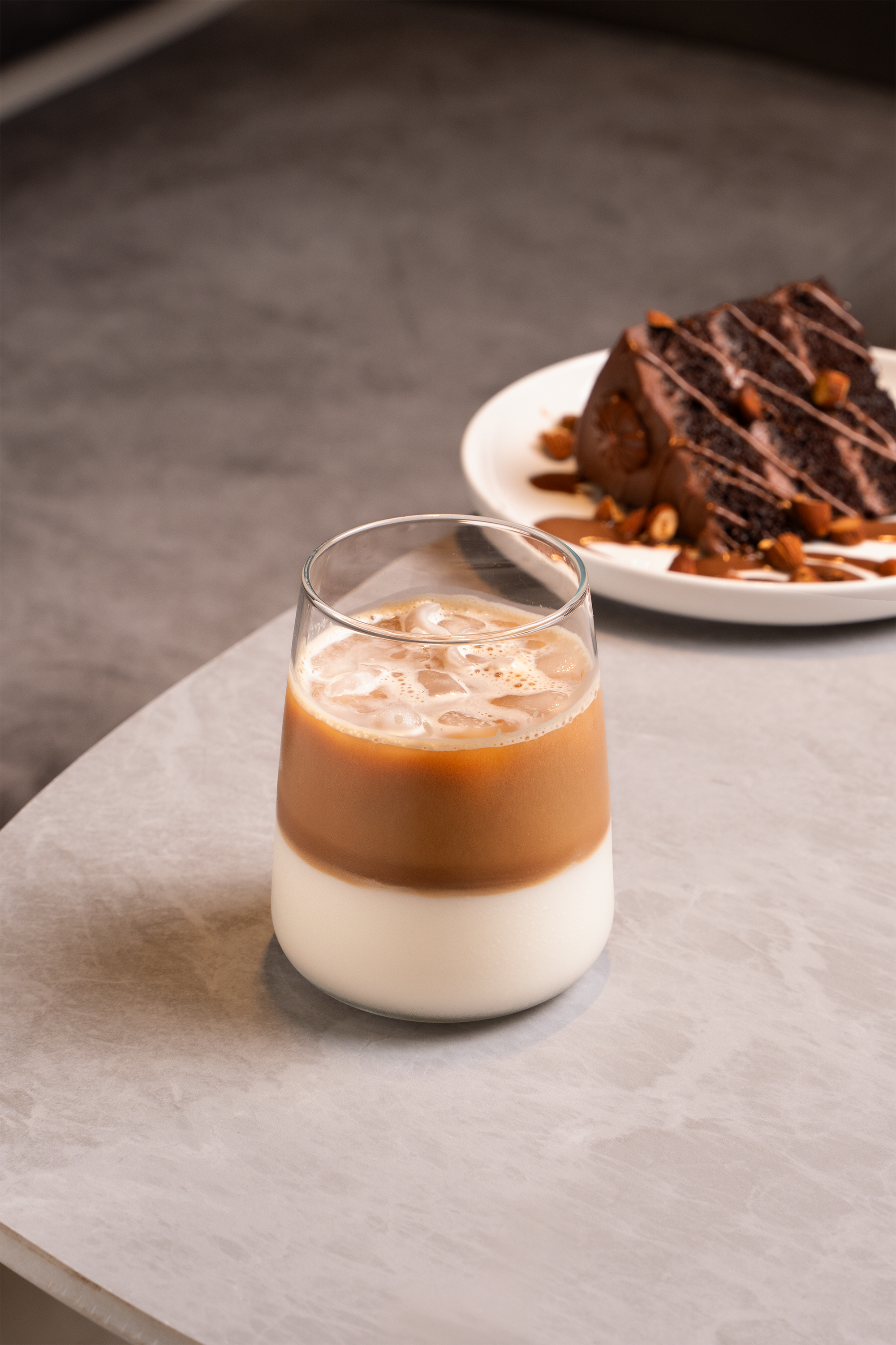 A glass of iced coffee with milk on a light-colored surface in front of a chocolate cake with nuts on a white plate.