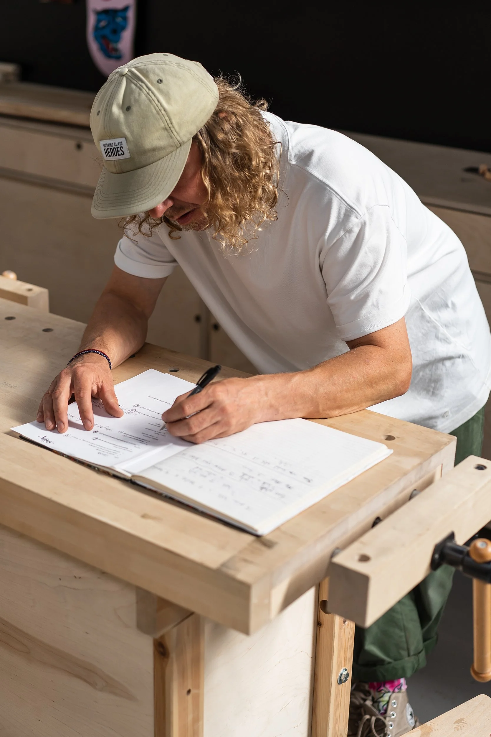 A person with curly hair and a beige cap leaning over a wooden workbench, writing in a notebook and working on documents in a workshop.