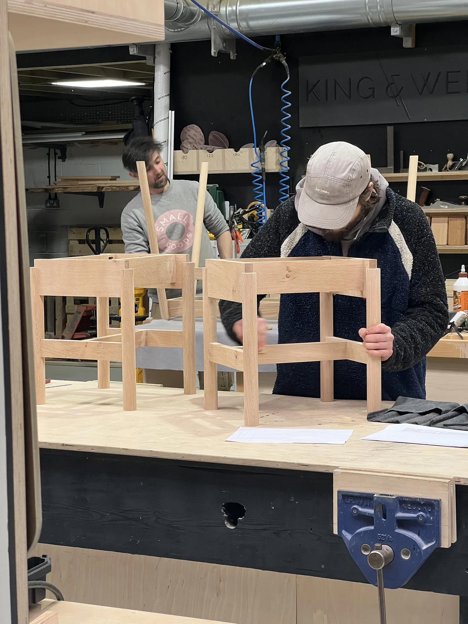 Two people working in a woodworking shop, assembling light-colored wooden chairs at a workbench. One person is focused on their task, wearing a baseball cap and a fleece jacket, while the other, in a gray hoodie, looks on.