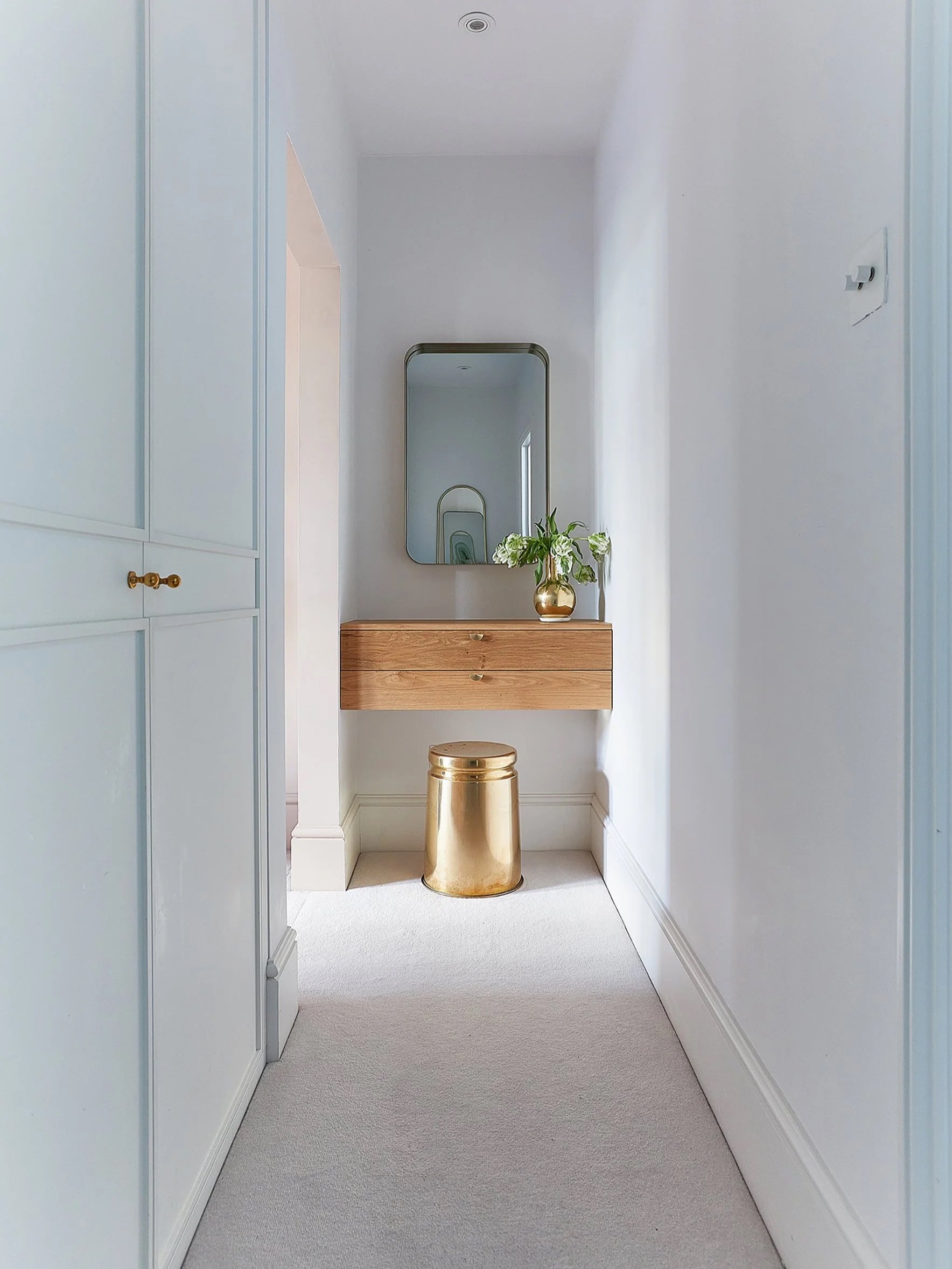 A narrow hallway with white walls and light-colored carpet, featuring a wooden wall-mounted console, a large mirror, a gold vase with flowers, and a gold stool.