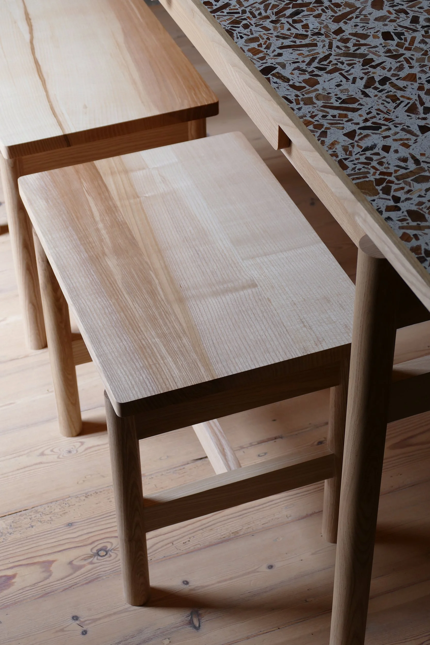 Wooden furniture including two stools and part of a table with a terrazzo-style top in a room with wooden flooring.