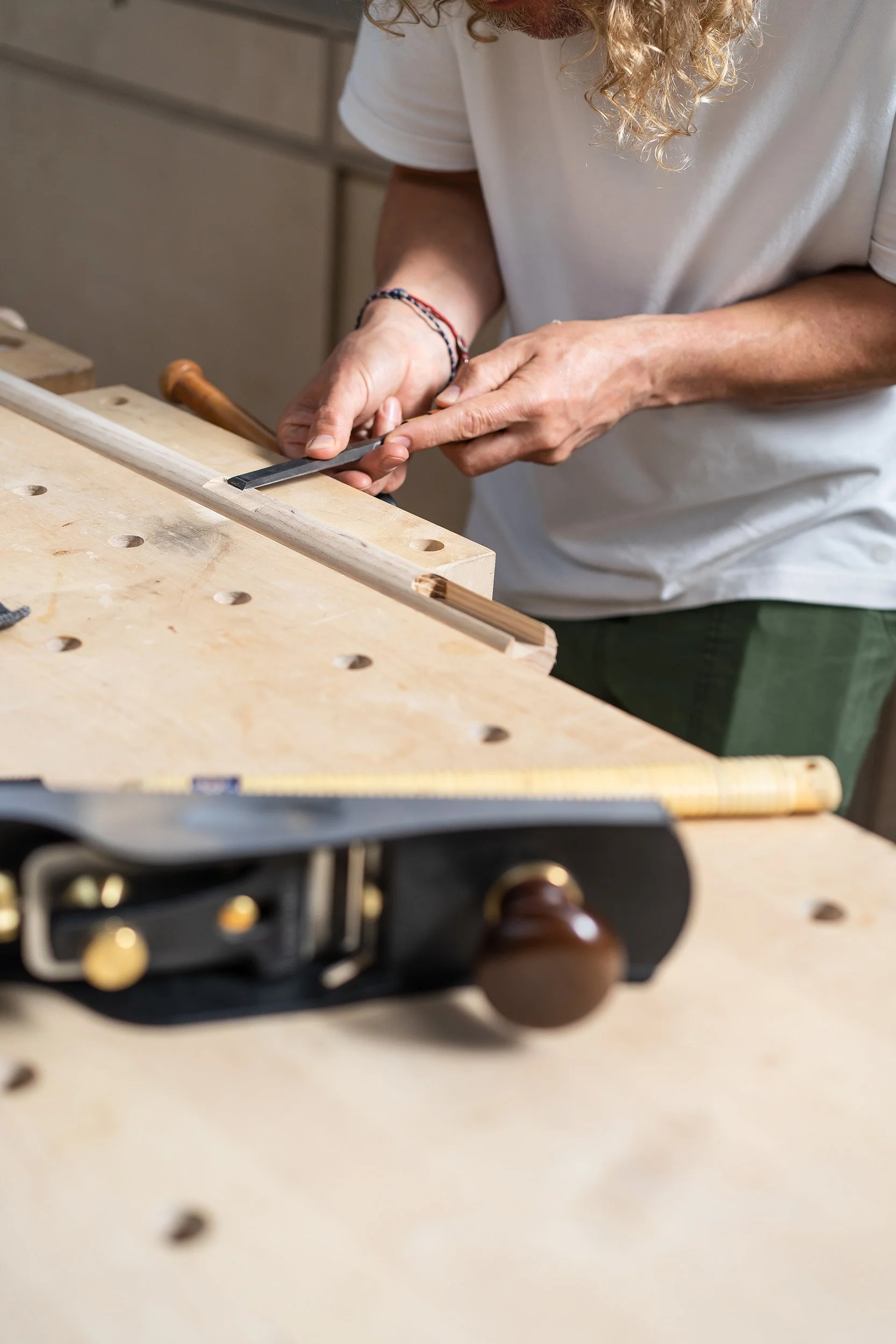 A person with curly blonde hair and wearing a white t-shirt is using a chisel and hammer to carve a piece of wood on a workbench.