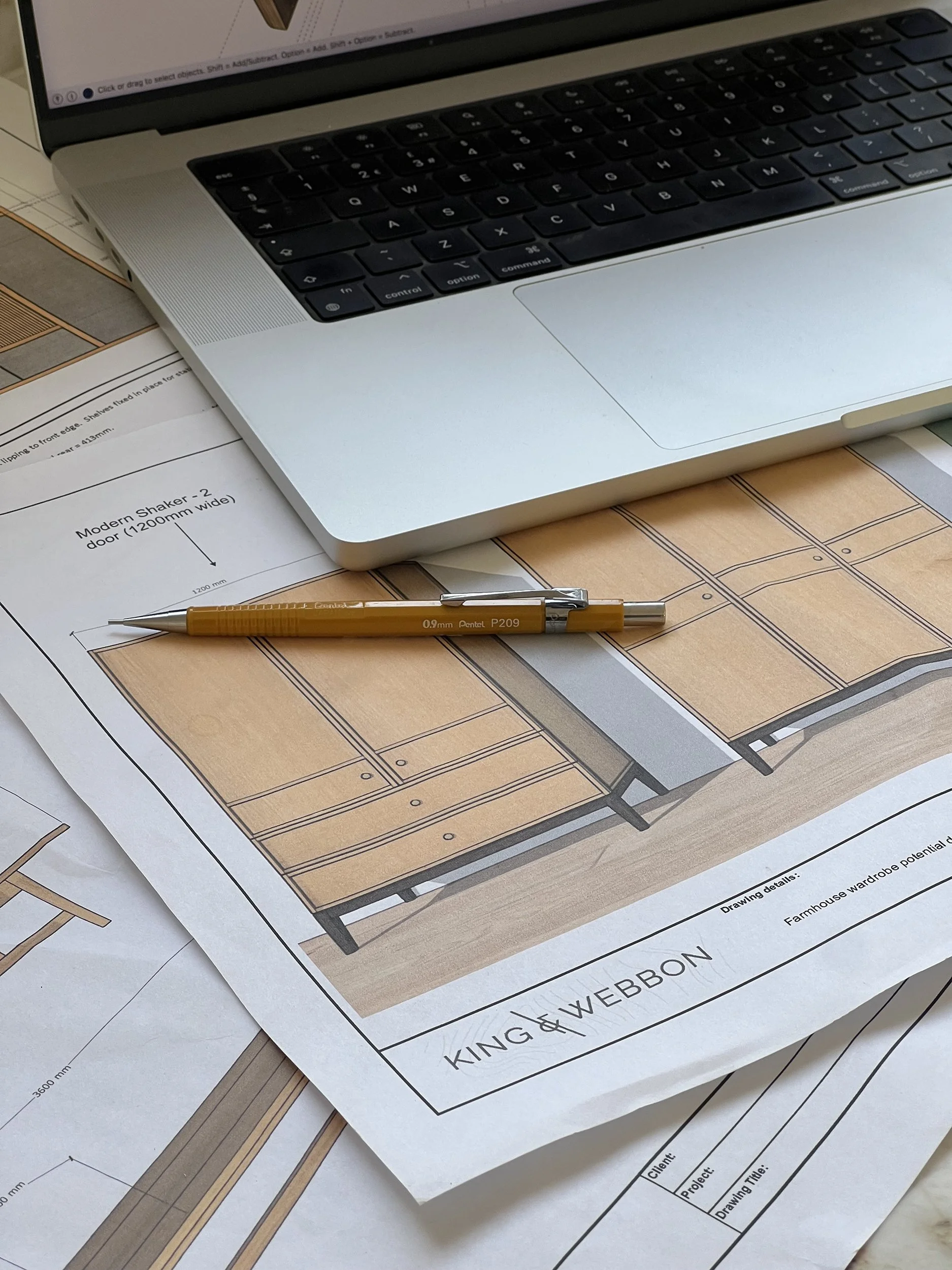 Architectural drawings and plans for a kitchen and wardrobe design, with a yellow pen and a partially open laptop on top of the plans.