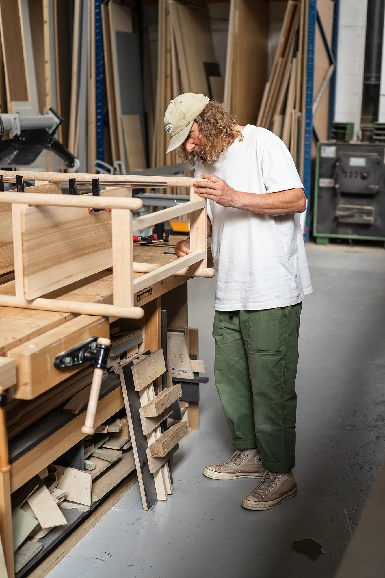 A man in a white t-shirt, green pants, and tan sneakers is working on a woodworking project in a workshop. He is holding a piece of wood and examining it carefully, with various tools and wood pieces on the workbench.