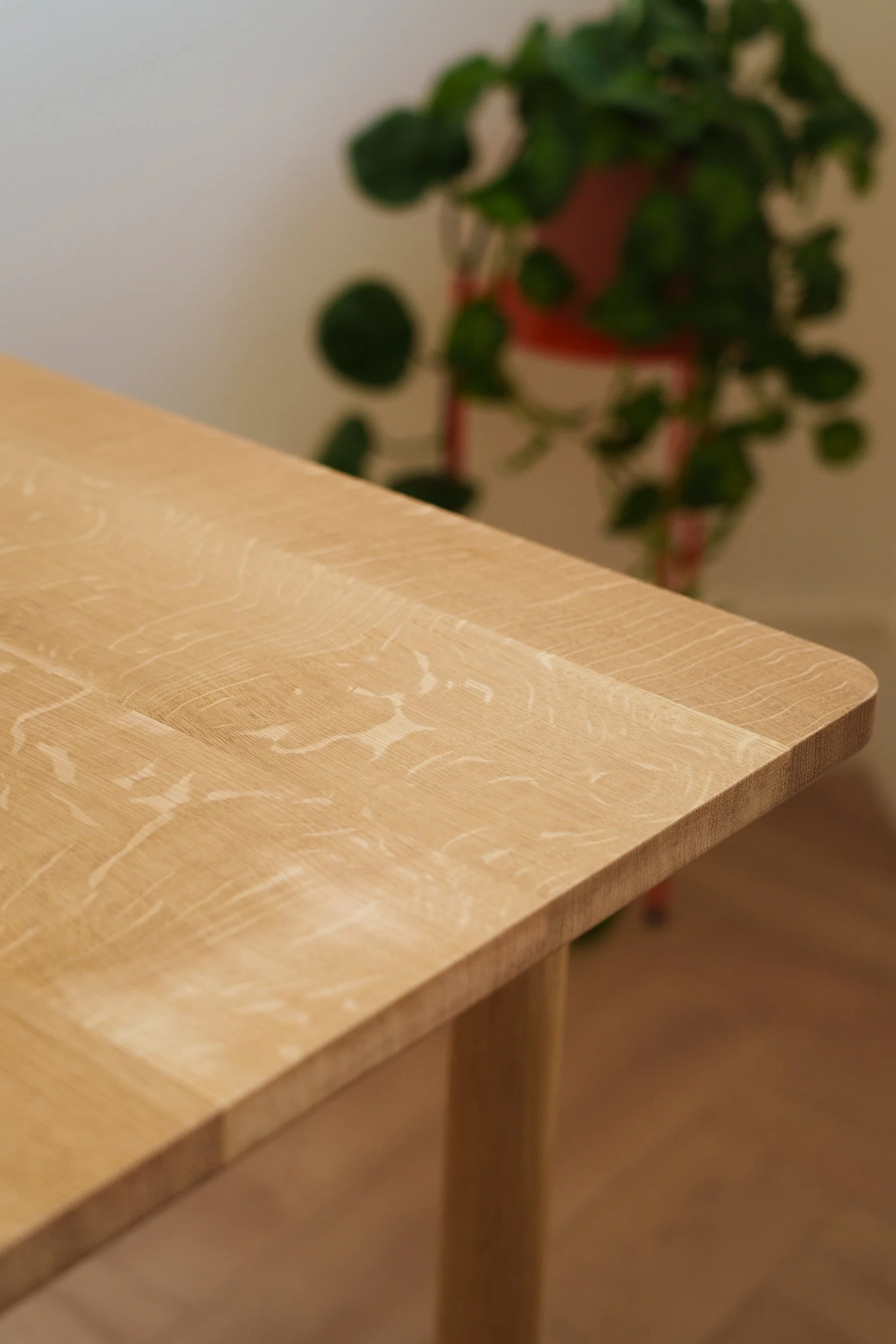 Close-up of a light wooden table with a blurred green potted plant in the background.