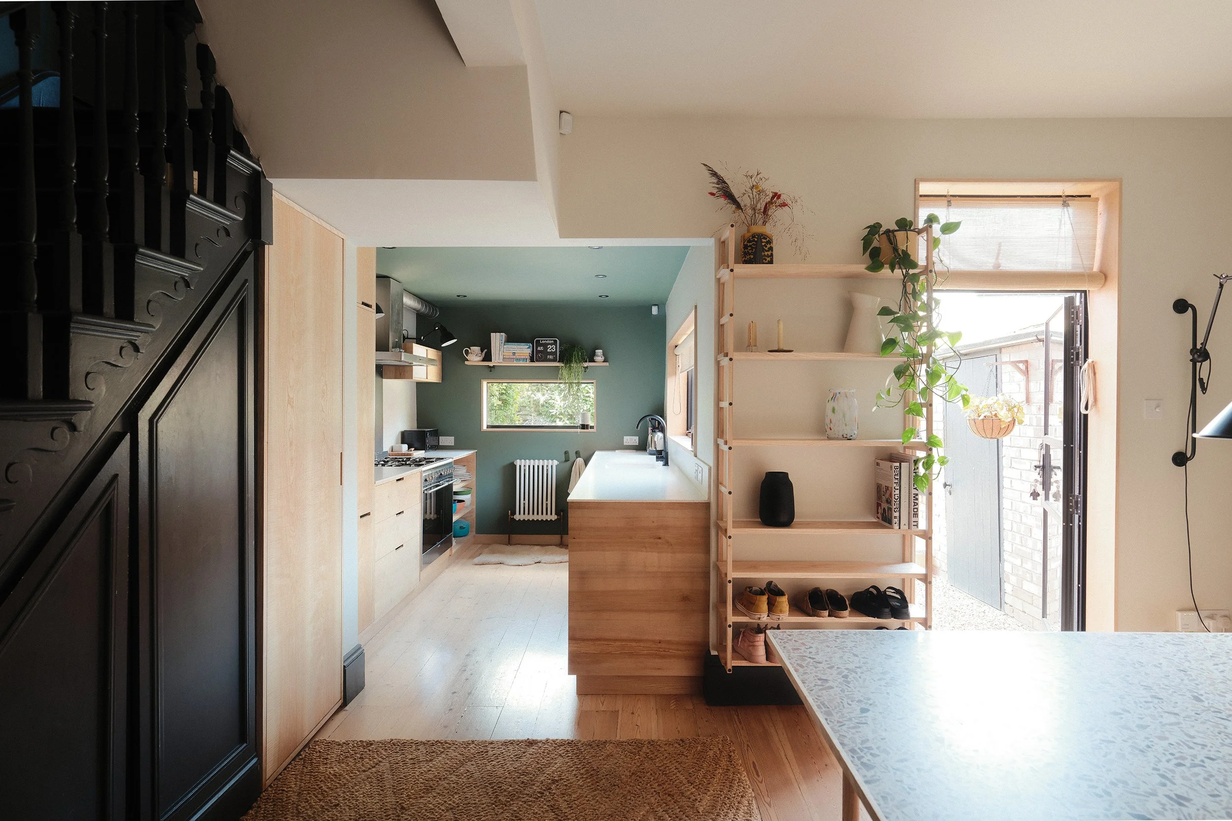 Bright, modern kitchen with wooden cabinetry, green accent wall, and large window, adjacent to an open living space with a wooden shelf and a sliding glass door leading outside.
