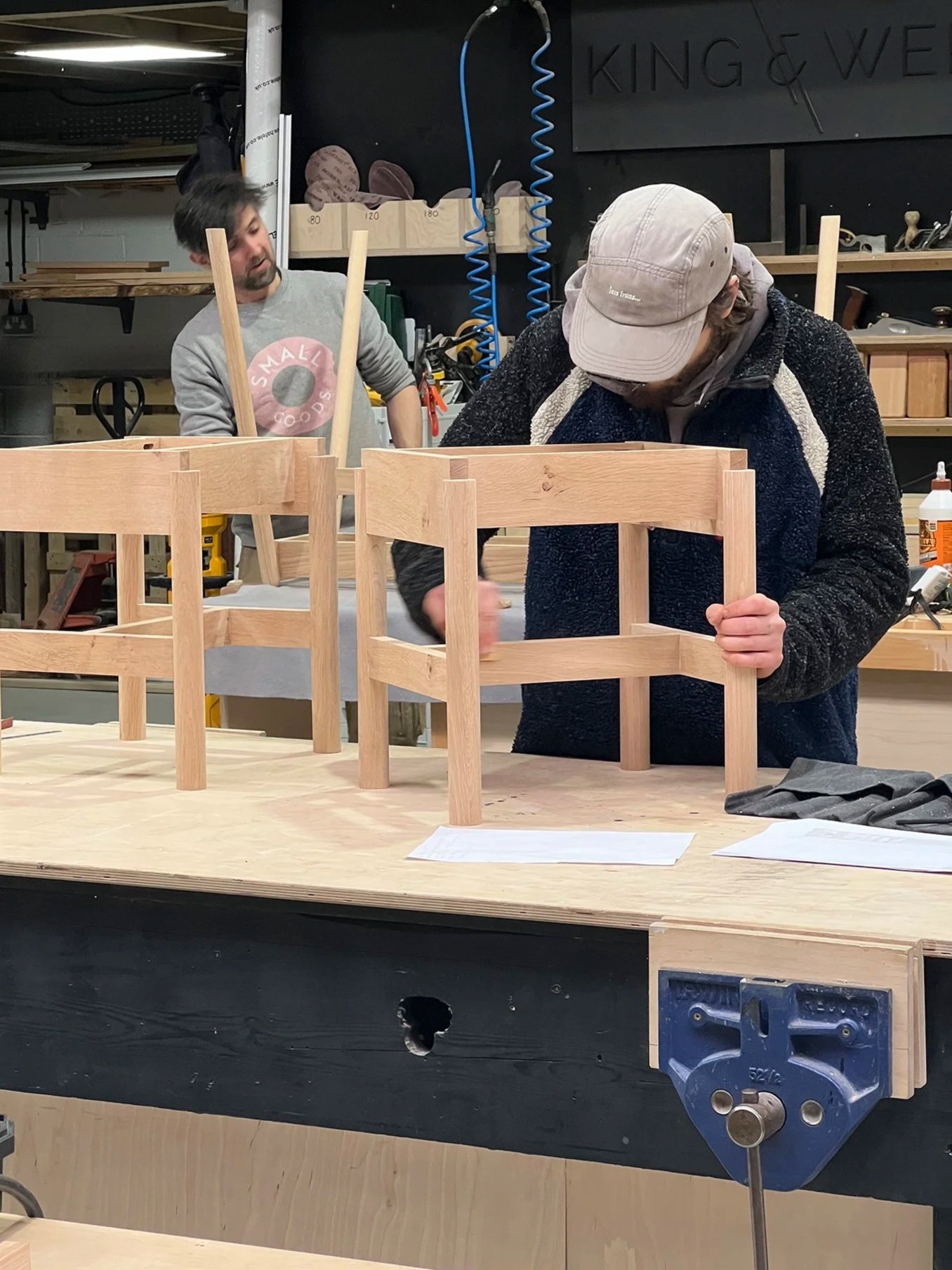 Two men working on assembling wooden furniture in a workshop, with one tightening screws and the other observing. Various tools and supplies are visible in the background.