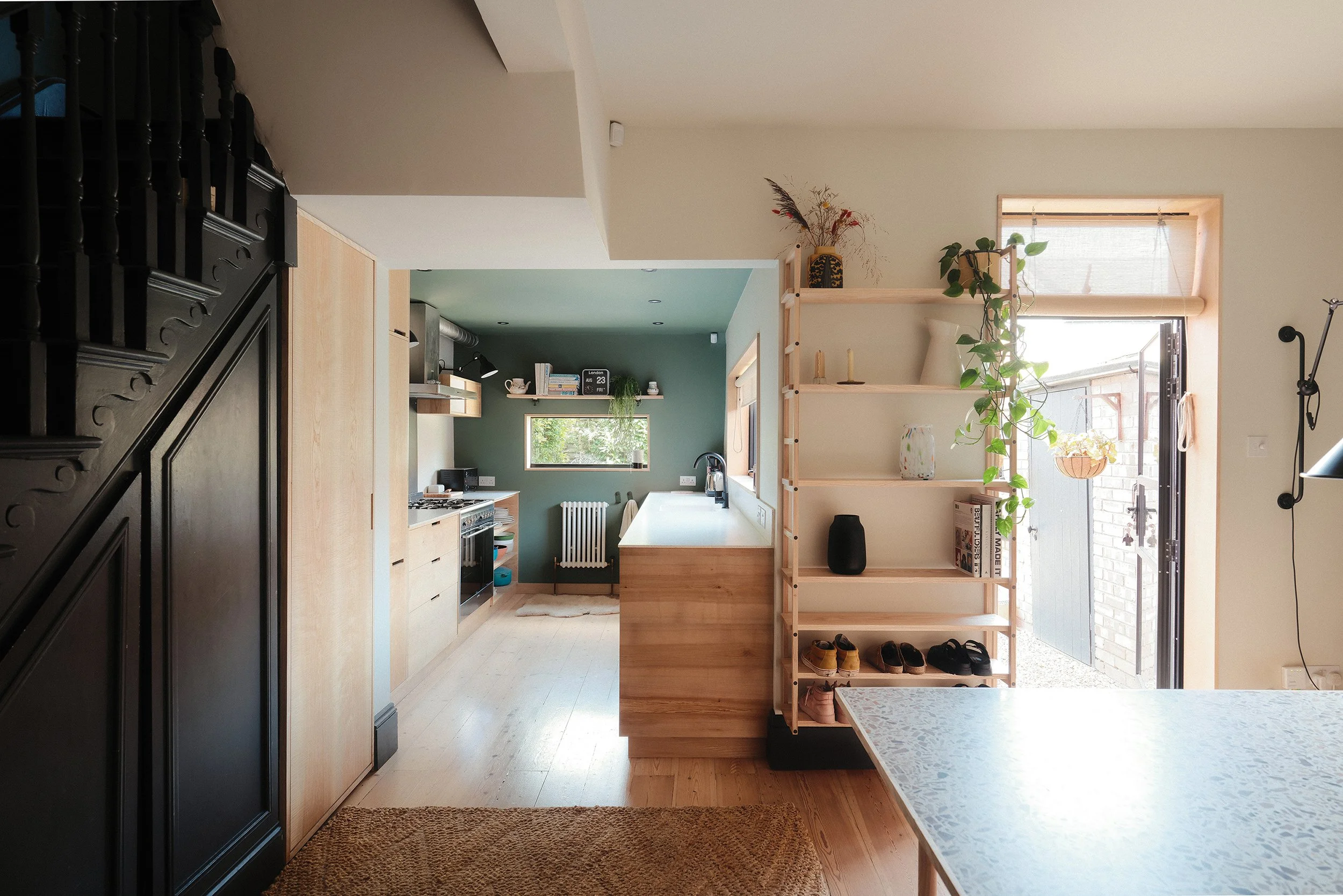Interior of a modern kitchen and dining area with light wood cabinets, green workspace wall, and open shelving, natural light, plants, and shoes near outdoor door.