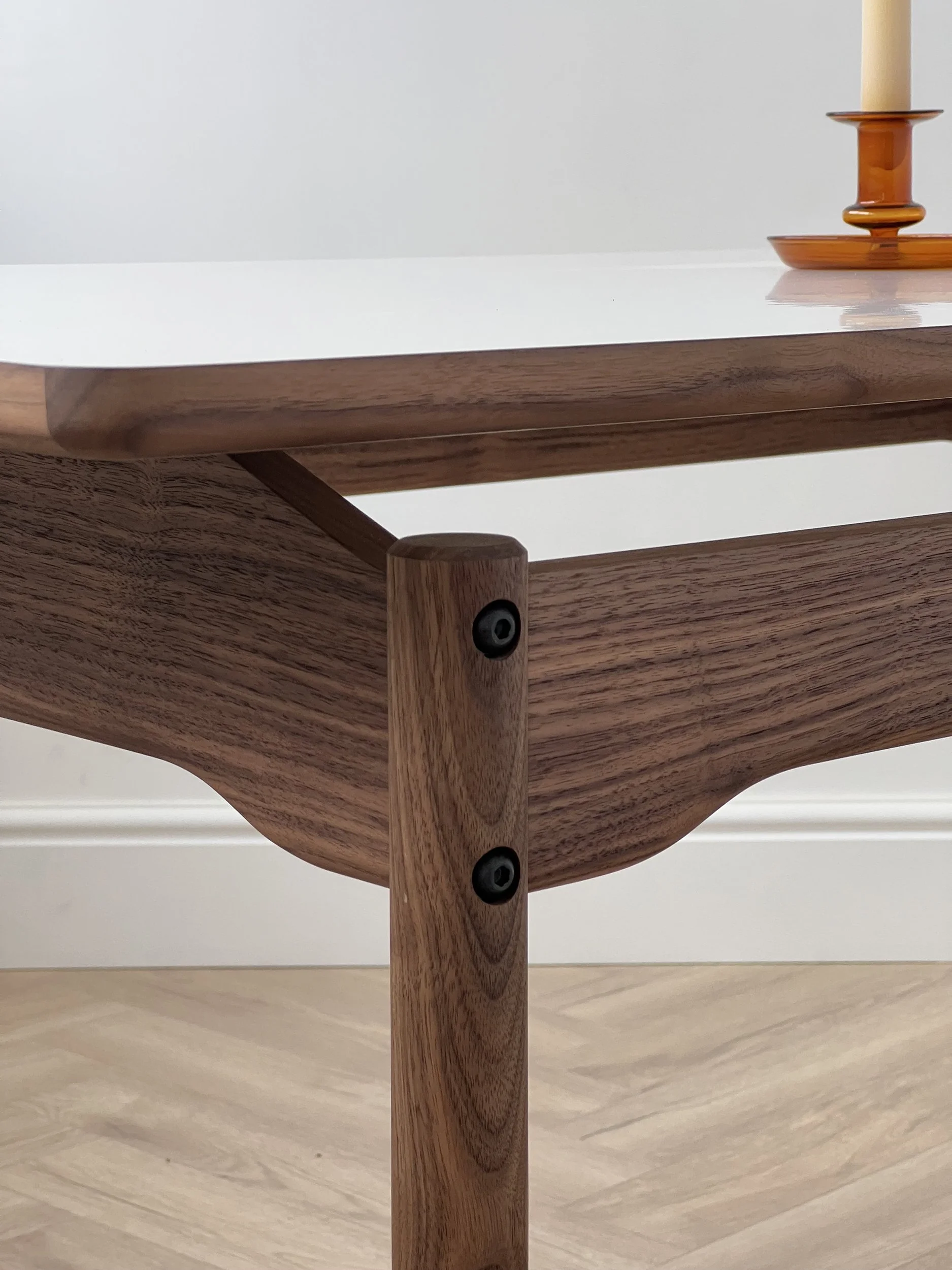 Close-up of a wooden table corner with black screws and a white tabletop, with a candle holder on the top right.