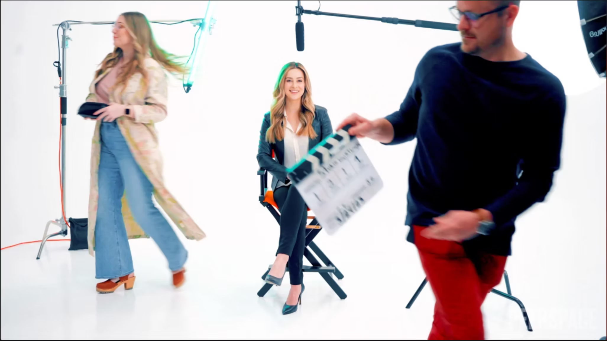 Film crew in a white studio capturing a woman sitting on a chair, a woman walking past, and a man holding a clapperboard with a microphone overhead.