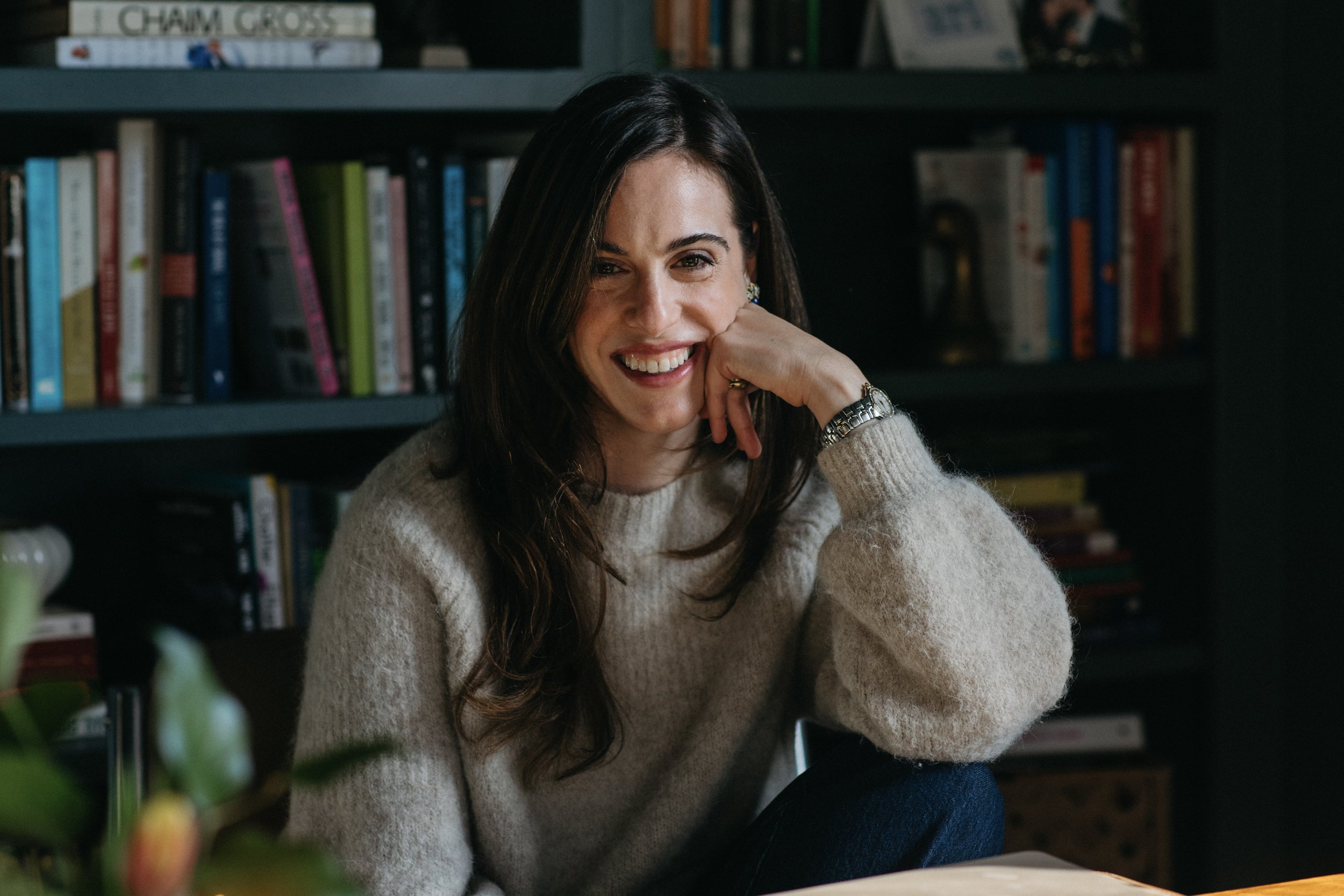 A woman with long dark hair, wearing a beige sweater, sitting indoors in front of a bookshelf filled with books, smiling and resting her head on her hand.