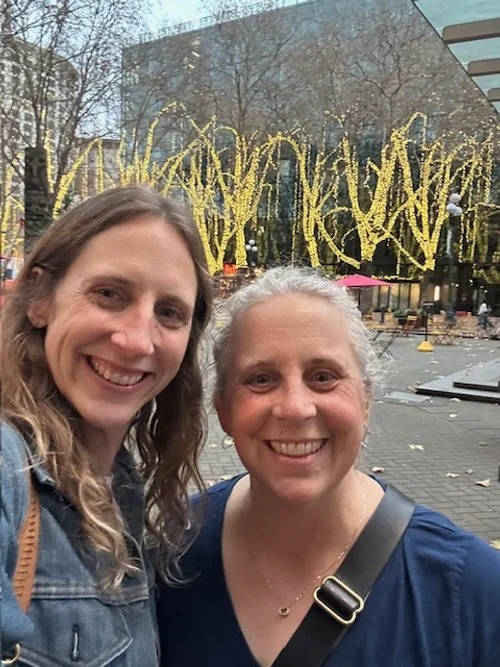 Two women smiling for a selfie outdoors with yellow string lights and a modern glass building in the background.
