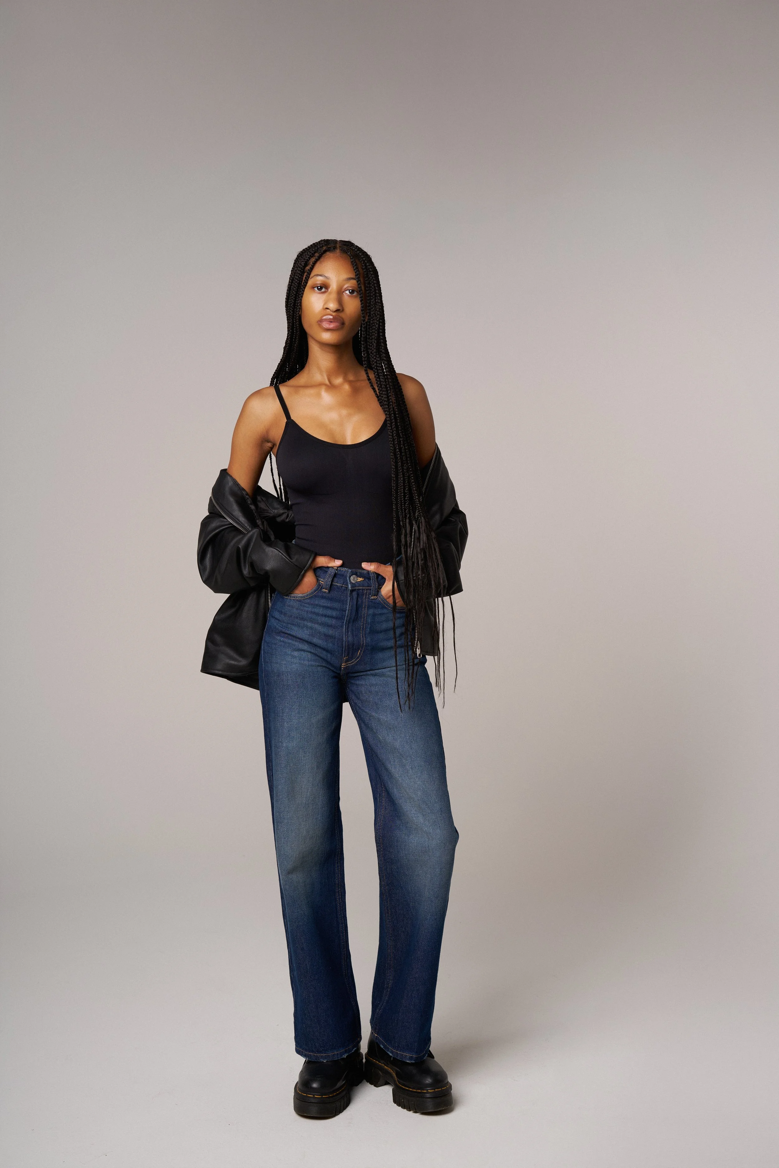 A woman with long braids wearing a black tank top, baggy blue jeans, black boots, and a black leather jacket draped over her shoulders, standing against a plain light gray background.