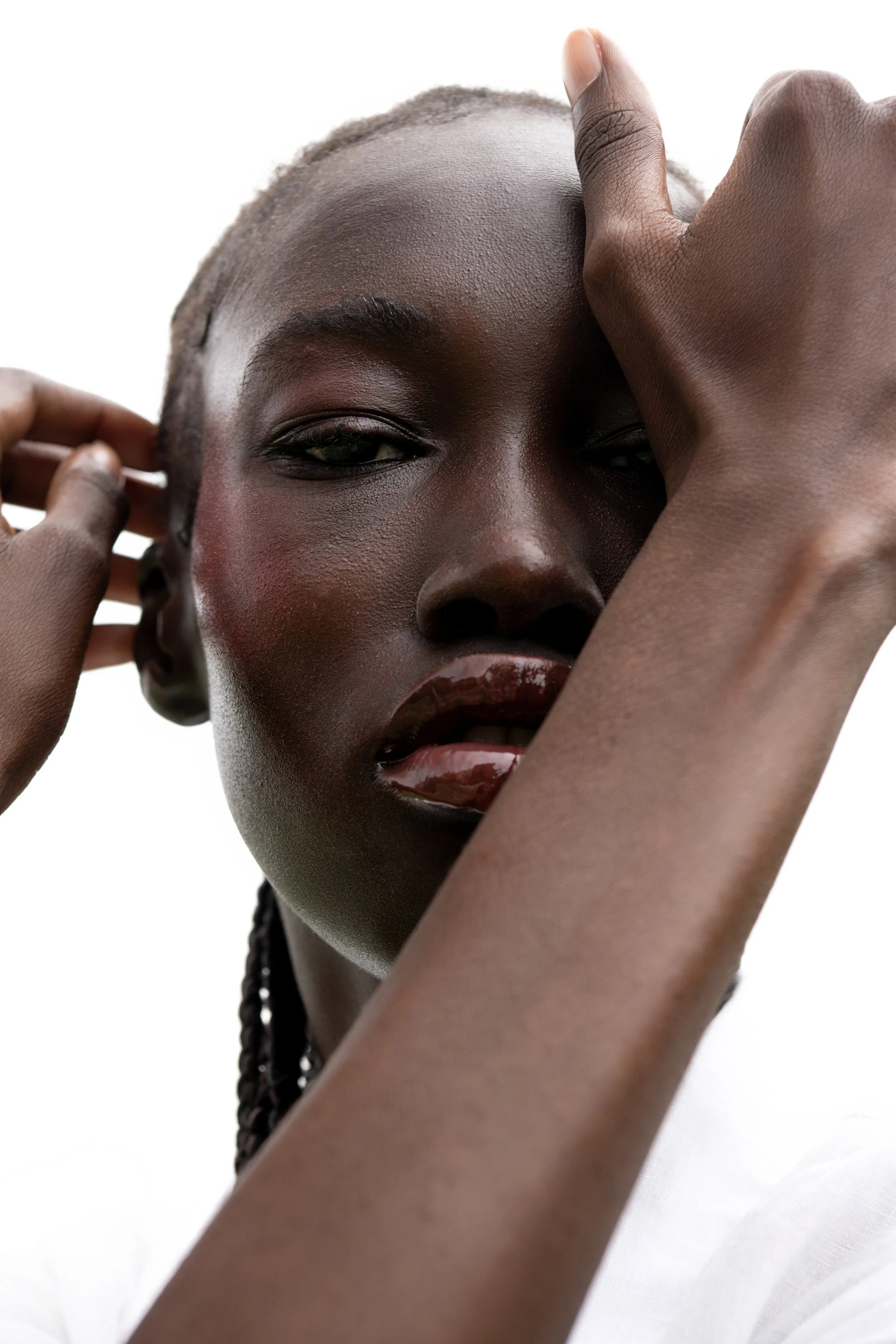 A close-up portrait of a Black woman with short hair, wearing shiny lipstick, with her hands touching her face and ears, against a white background.