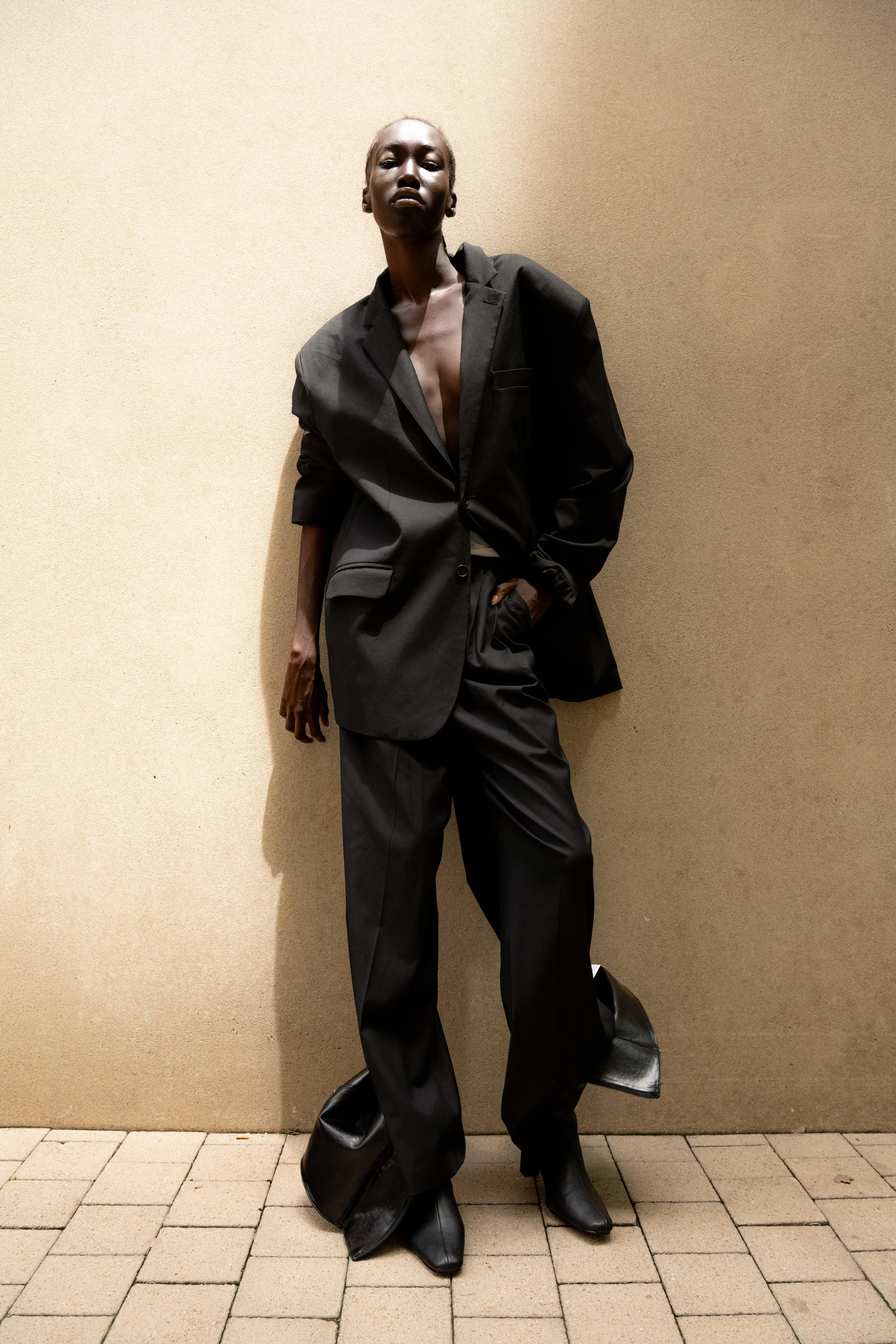 A person wearing oversized black suit with wide pants and a blazer, with their hand in their pocket, standing against a beige wall and looking at the camera.