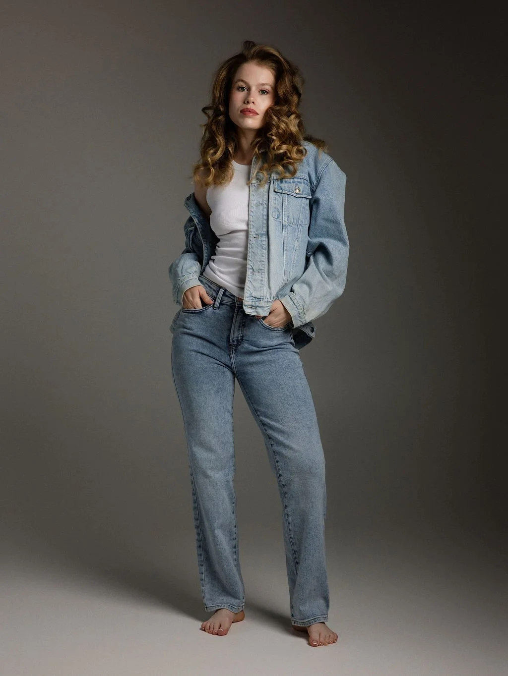 A woman with curly brown hair wearing a light blue denim jacket, white t-shirt, and high-waisted jeans, standing barefoot against a neutral background.