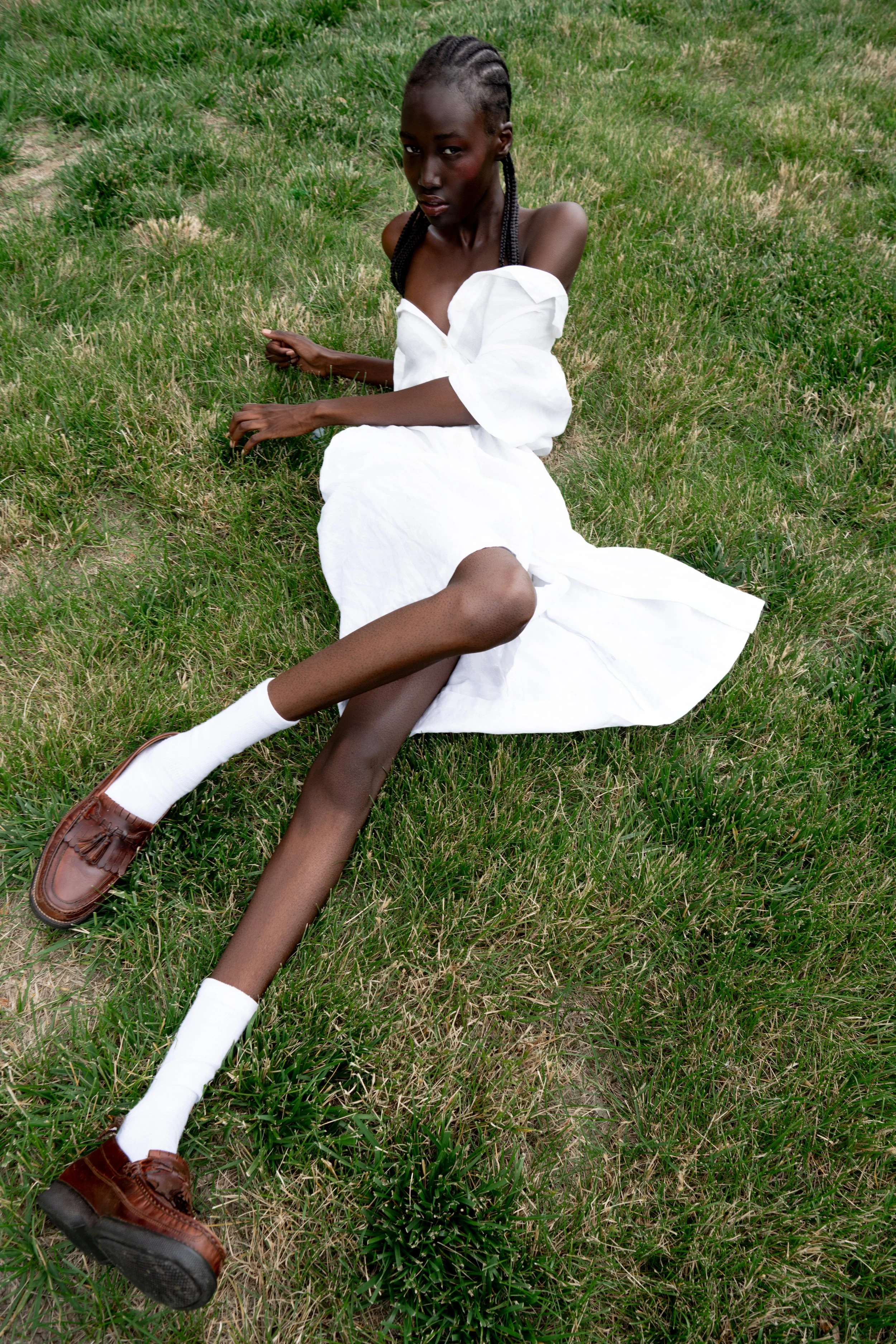 A woman with dark skin and braided hair sitting on green grass, wearing a white off-shoulder dress, white socks, and brown loafers, with a serious expression.