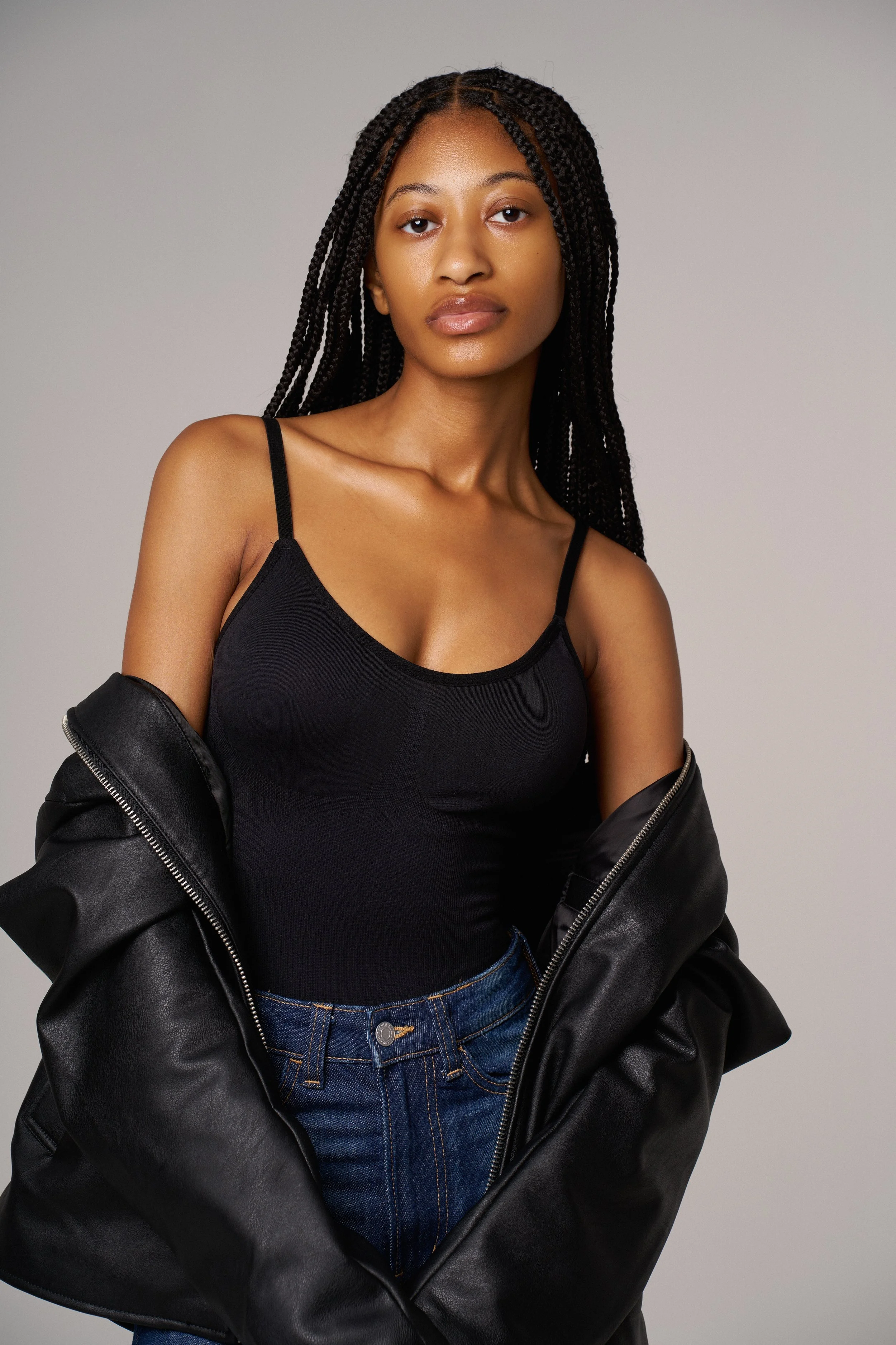 A woman with braided hair wearing a black spaghetti strap top, blue jeans, and a black leather jacket draped over her arms, standing against a plain gray background.