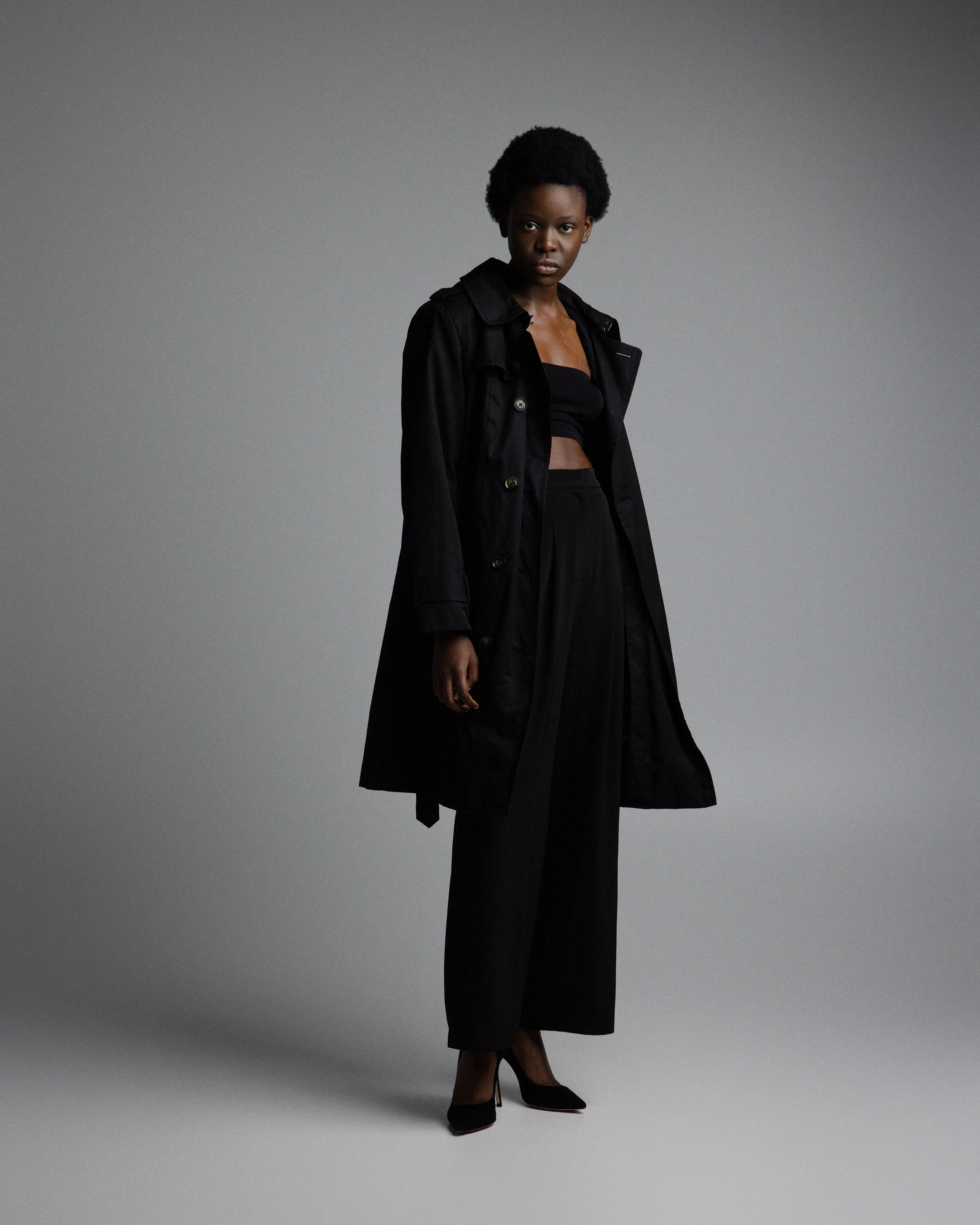 A woman with natural afro hairstyle wearing a black trench coat, black crop top, black wide-leg pants, and black high heels, standing against a plain gray background.