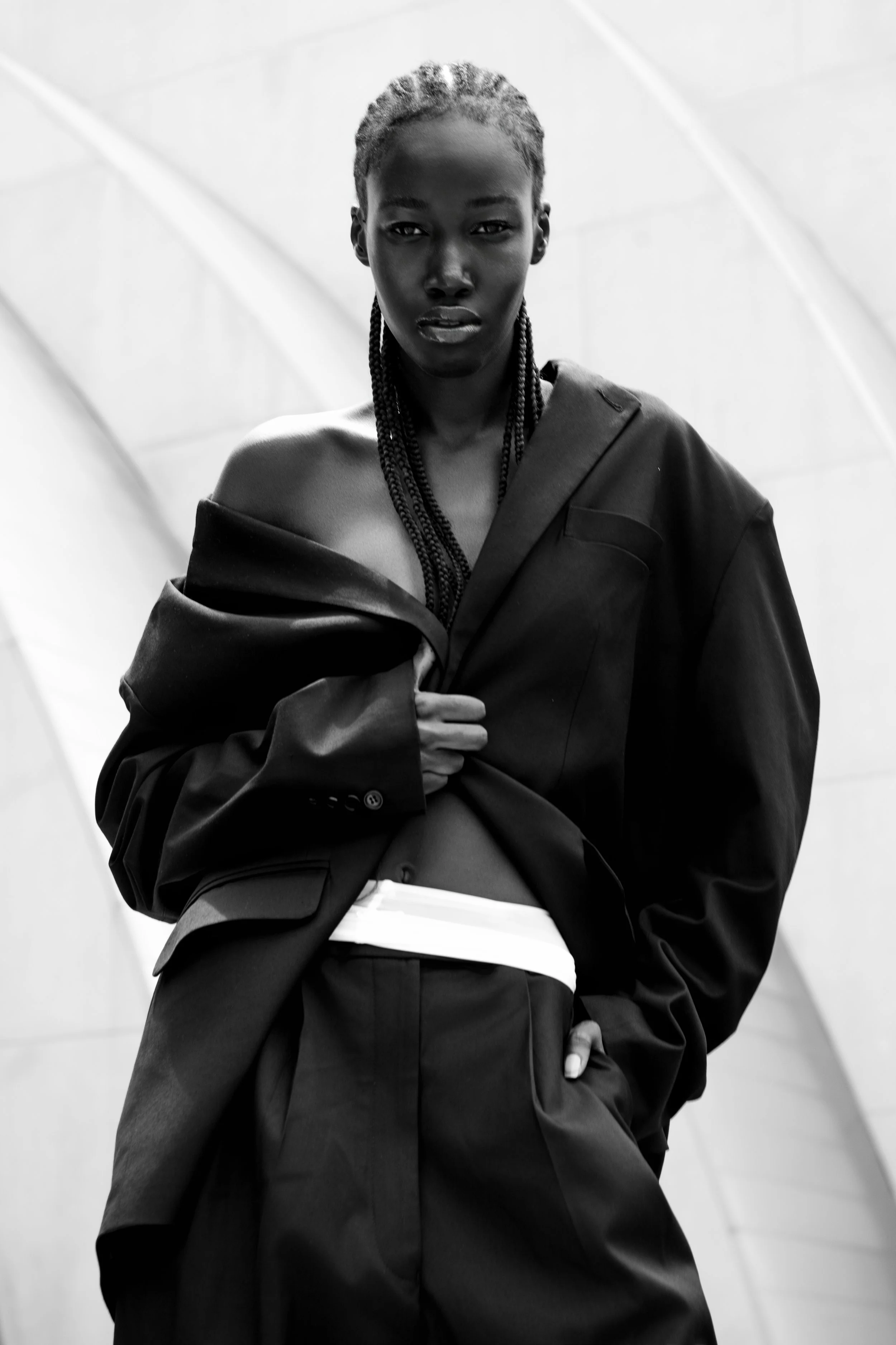 Black and white photo of a woman with braided hair wearing oversized blazer and pants, holding her open blazer to reveal her shoulder and midsection, with an abstract architectural background.