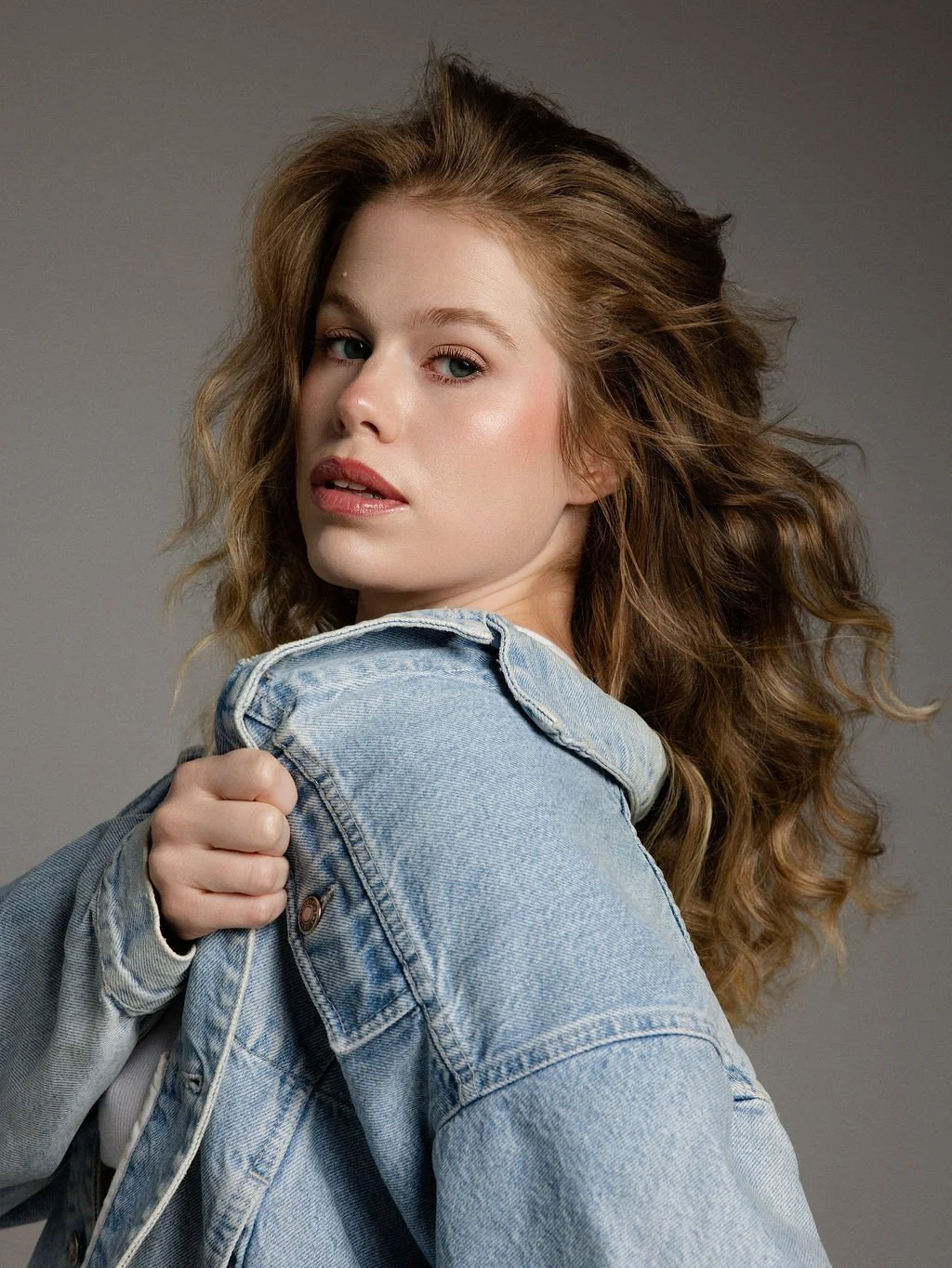 A young woman with long, curly auburn hair wearing a light blue denim jacket, looking over her shoulder against a plain gray background.
