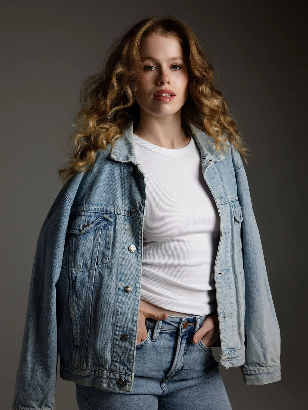 A young woman with curly red hair wearing a white t-shirt and a denim jacket, posing against a plain gray background.