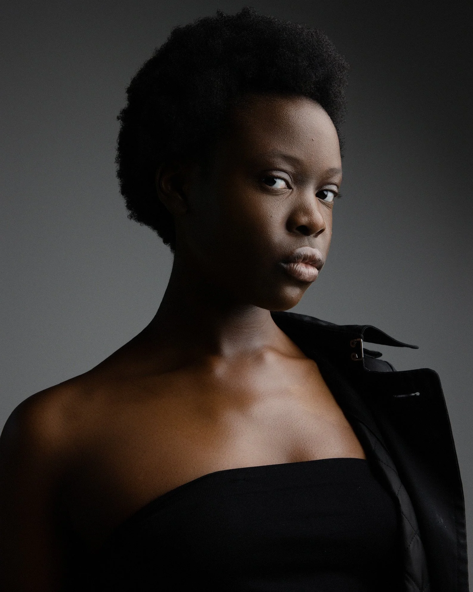Portrait of a young Black woman with natural hair, wearing a black strapless top and a black jacket, against a dark gray background.
