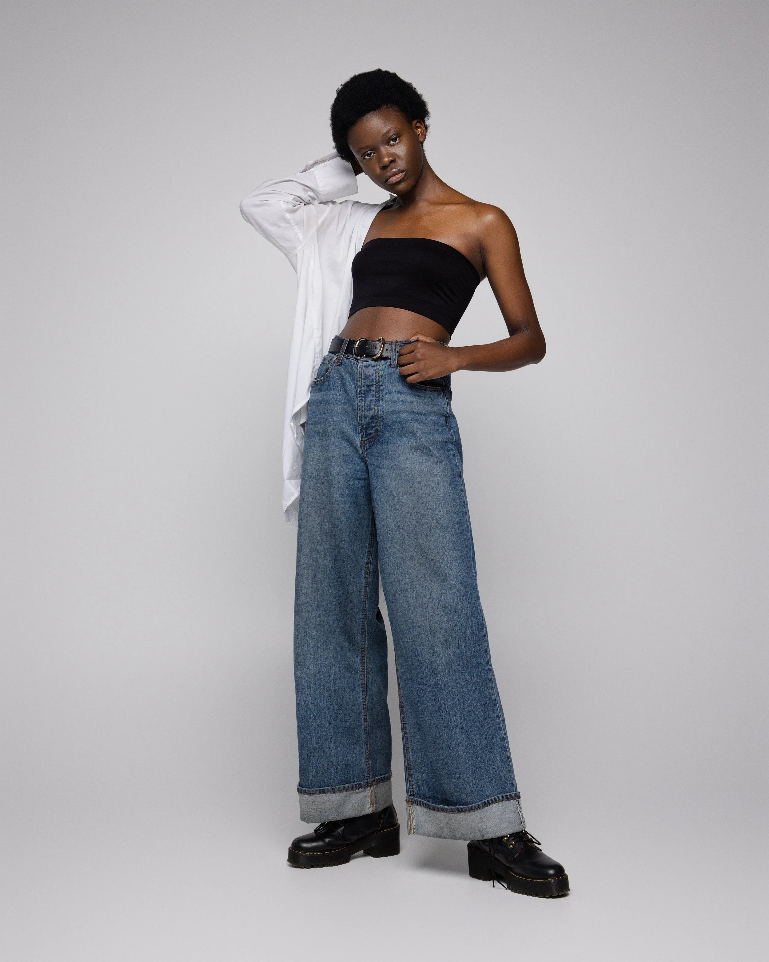 Young woman with short curly hair wearing a black strapless top, loose wide-leg jeans, black boots, and a partially unbuttoned white shirt over her shoulder, standing against a plain light gray background.