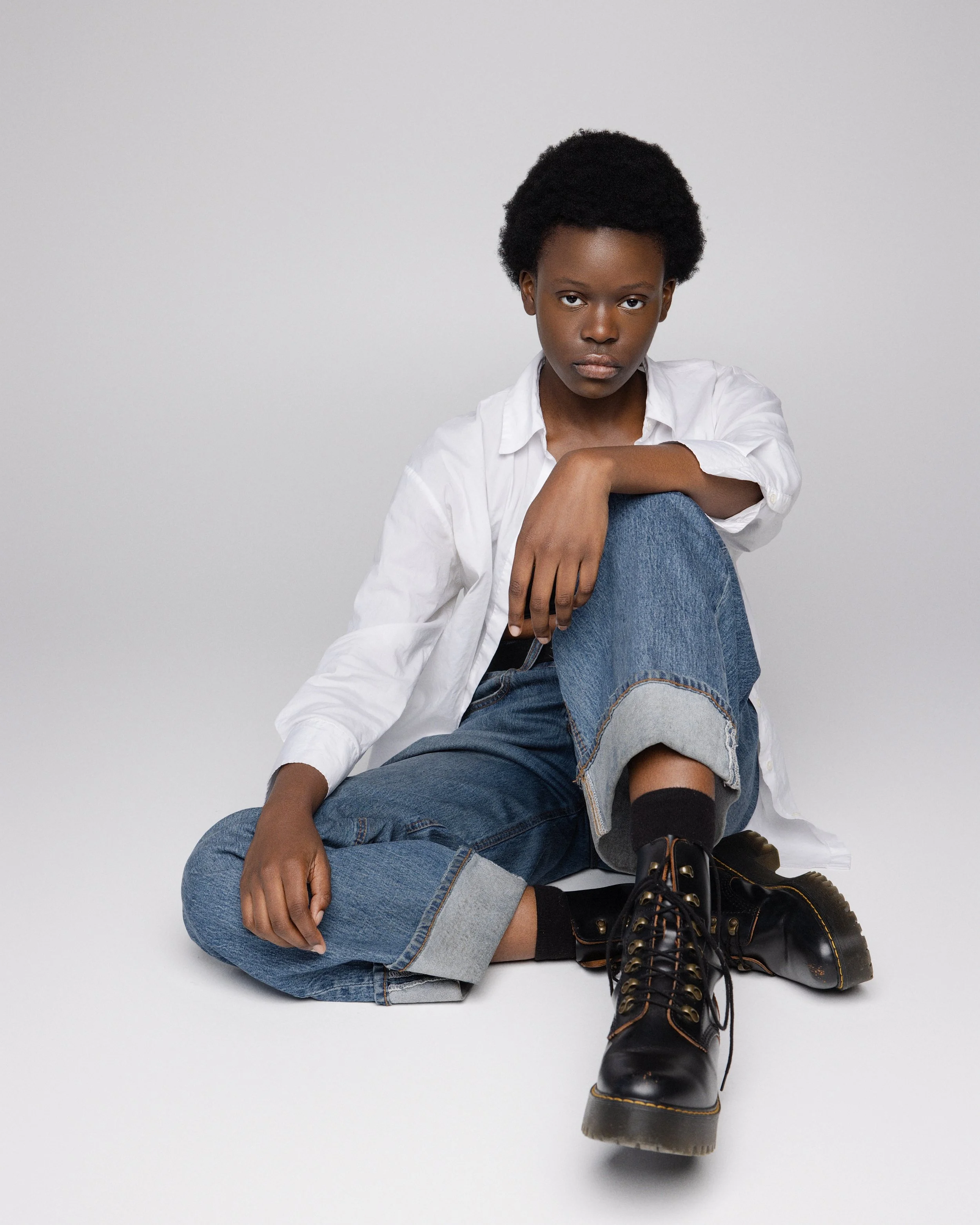 A young woman with short natural hair, wearing a white shirt, blue jeans, black boots, and black socks, sitting on the floor against a plain white background, looking directly at the camera with a serious expression.