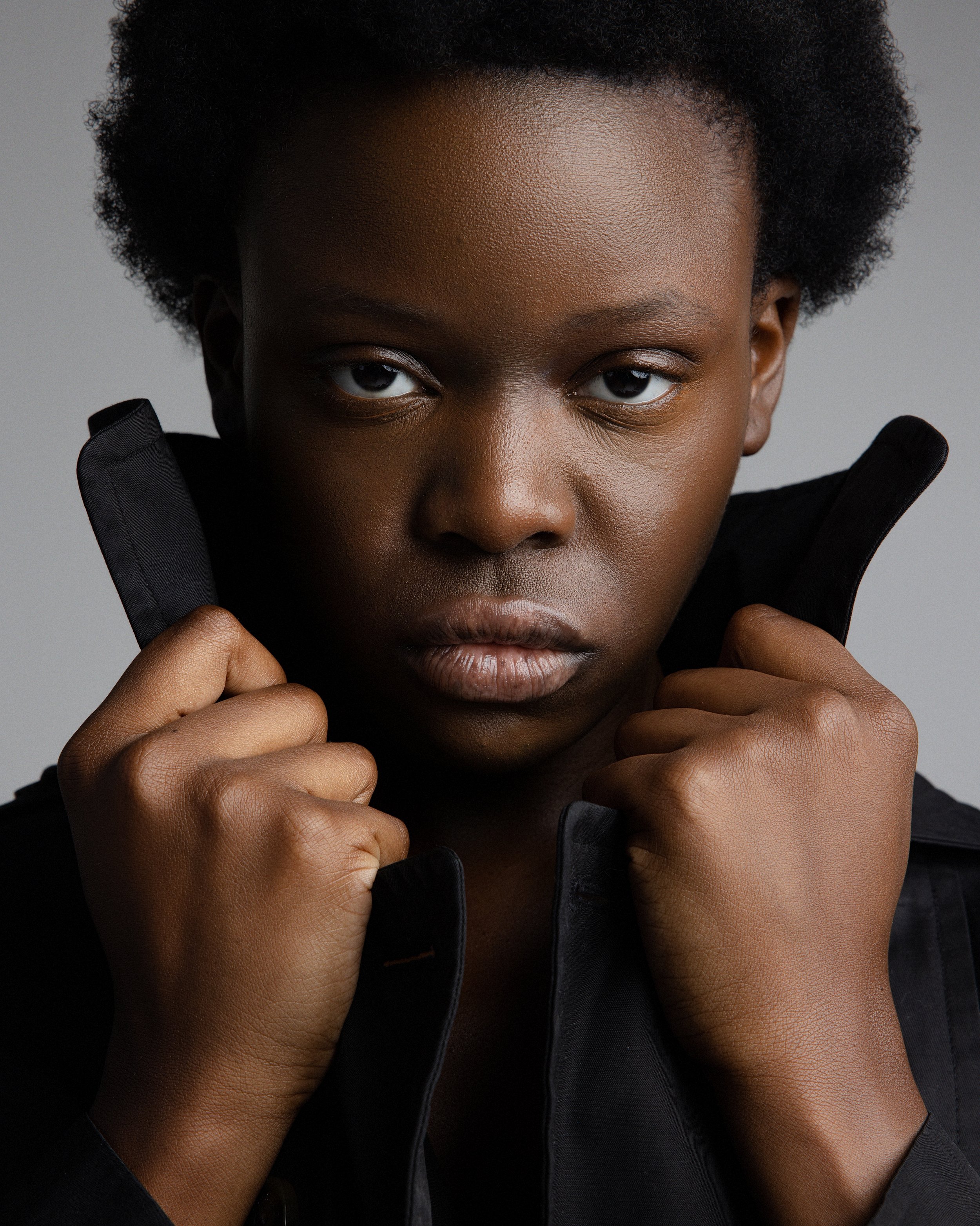 Close-up of a woman with natural hair wearing a black jacket, holding the collar with both hands, looking directly at the camera.