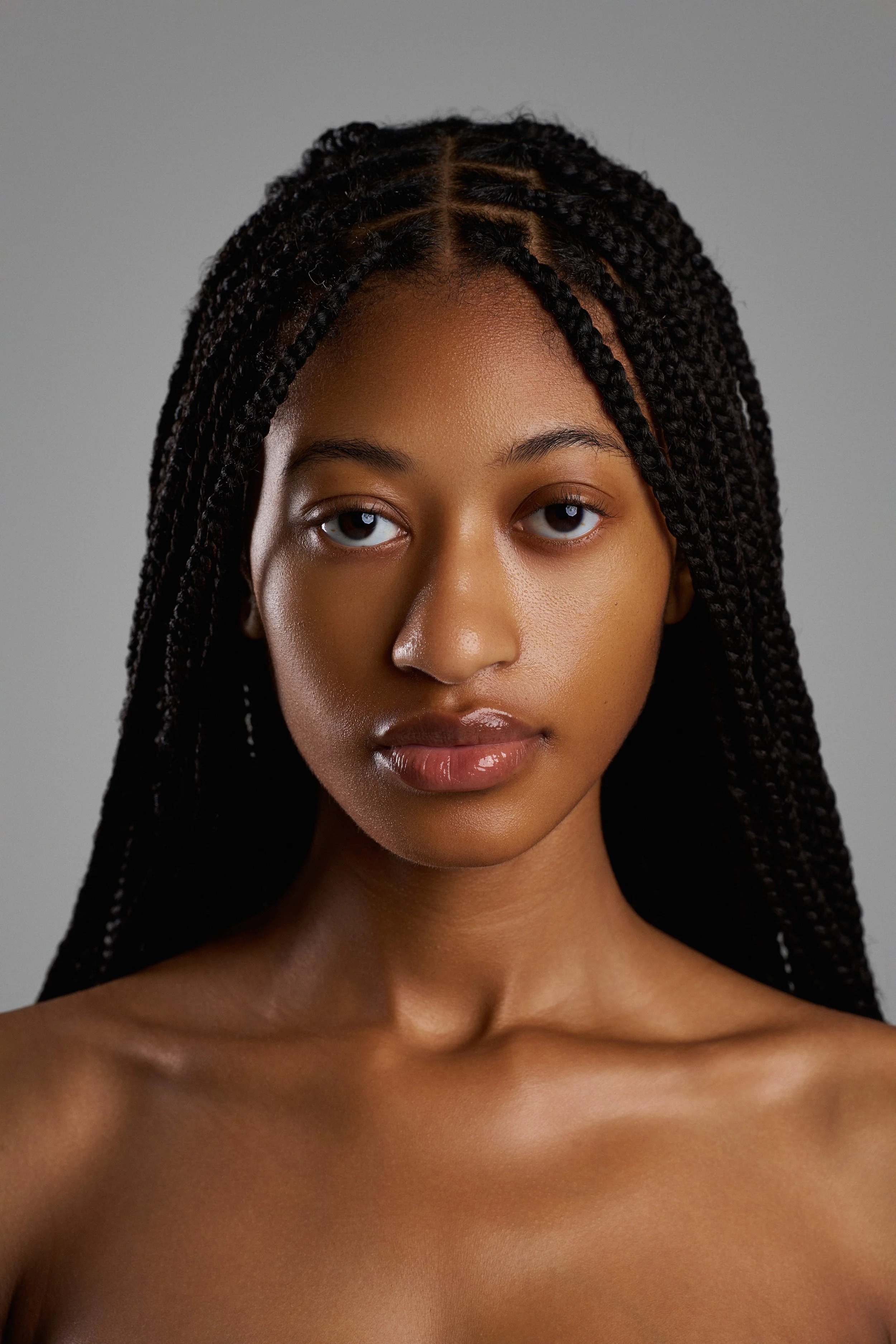 Close-up portrait of a young woman with long braided hair, looking directly at the camera with neutral expression, against a plain gray background.