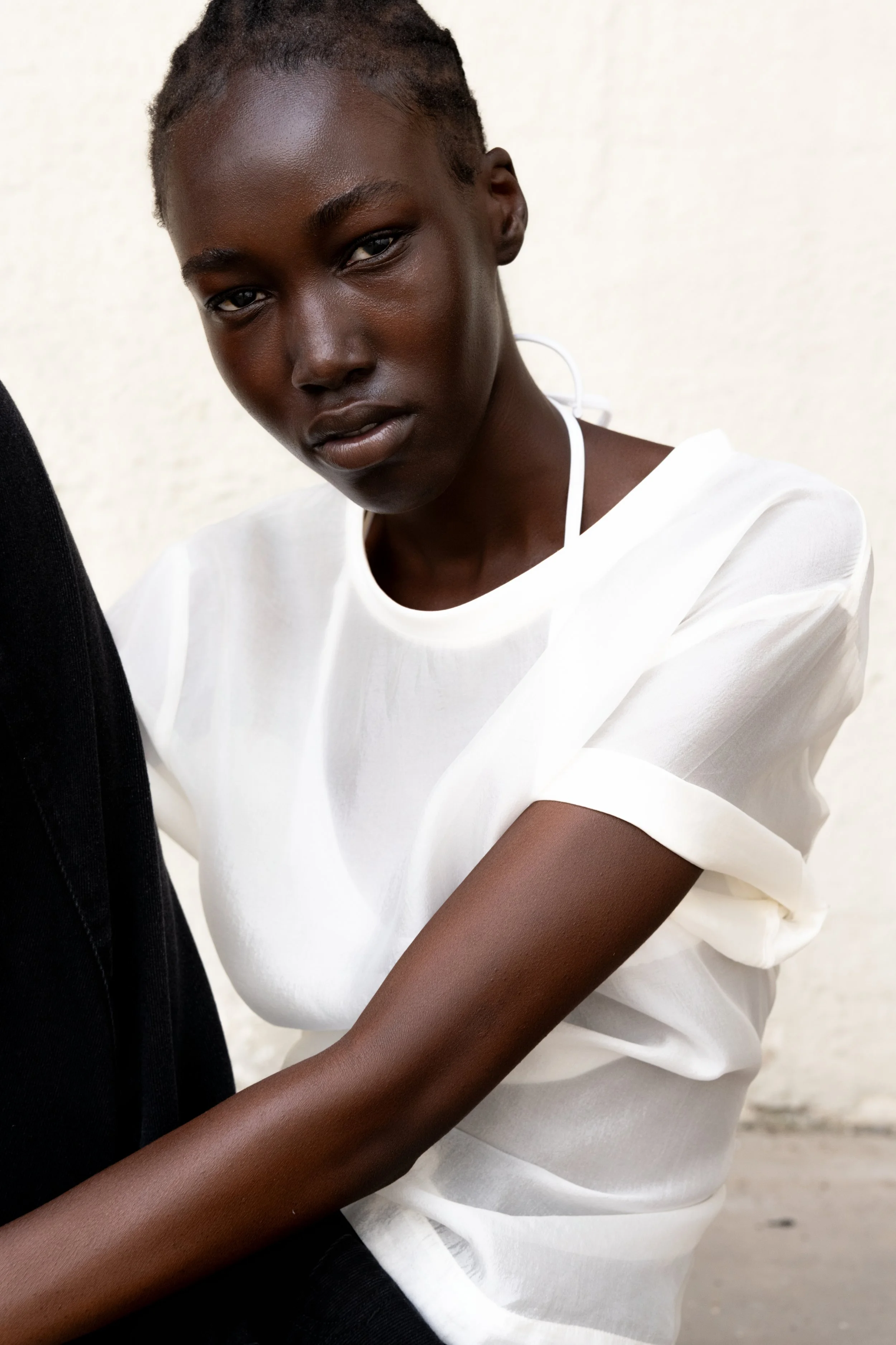 A young woman with dark skin and short dreadlocks, wearing a white top, looking directly at the camera against a neutral background.