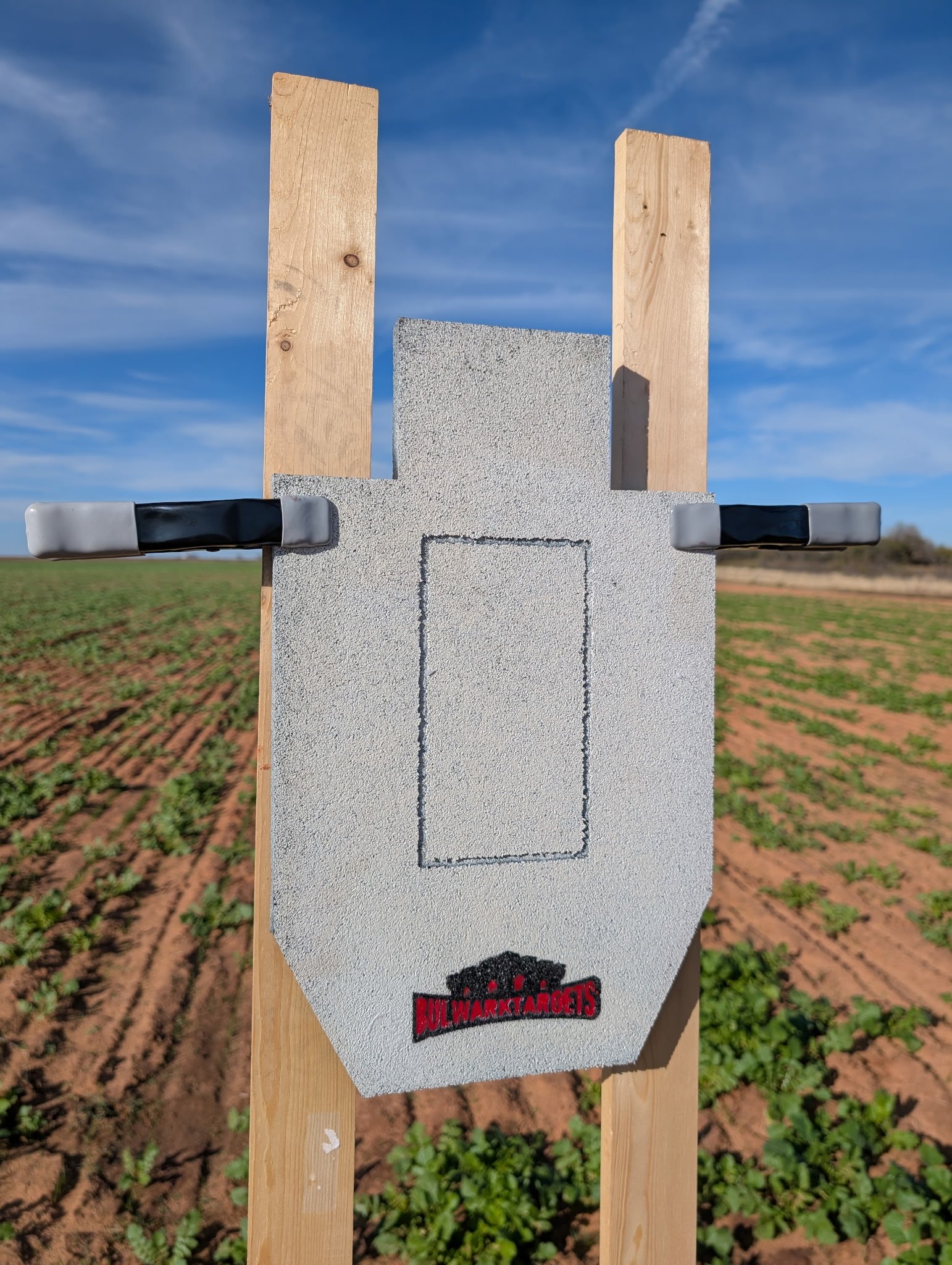 A white clay pigeon target with black and red logo, mounted on a wooden stand outdoors in a field with blue sky overhead.
