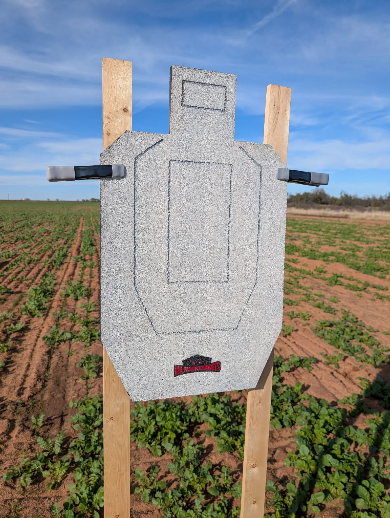 Outline of a bottle drawn on a foam board mounted on a wooden stake, set outdoors in a field with planted crops and a blue sky.