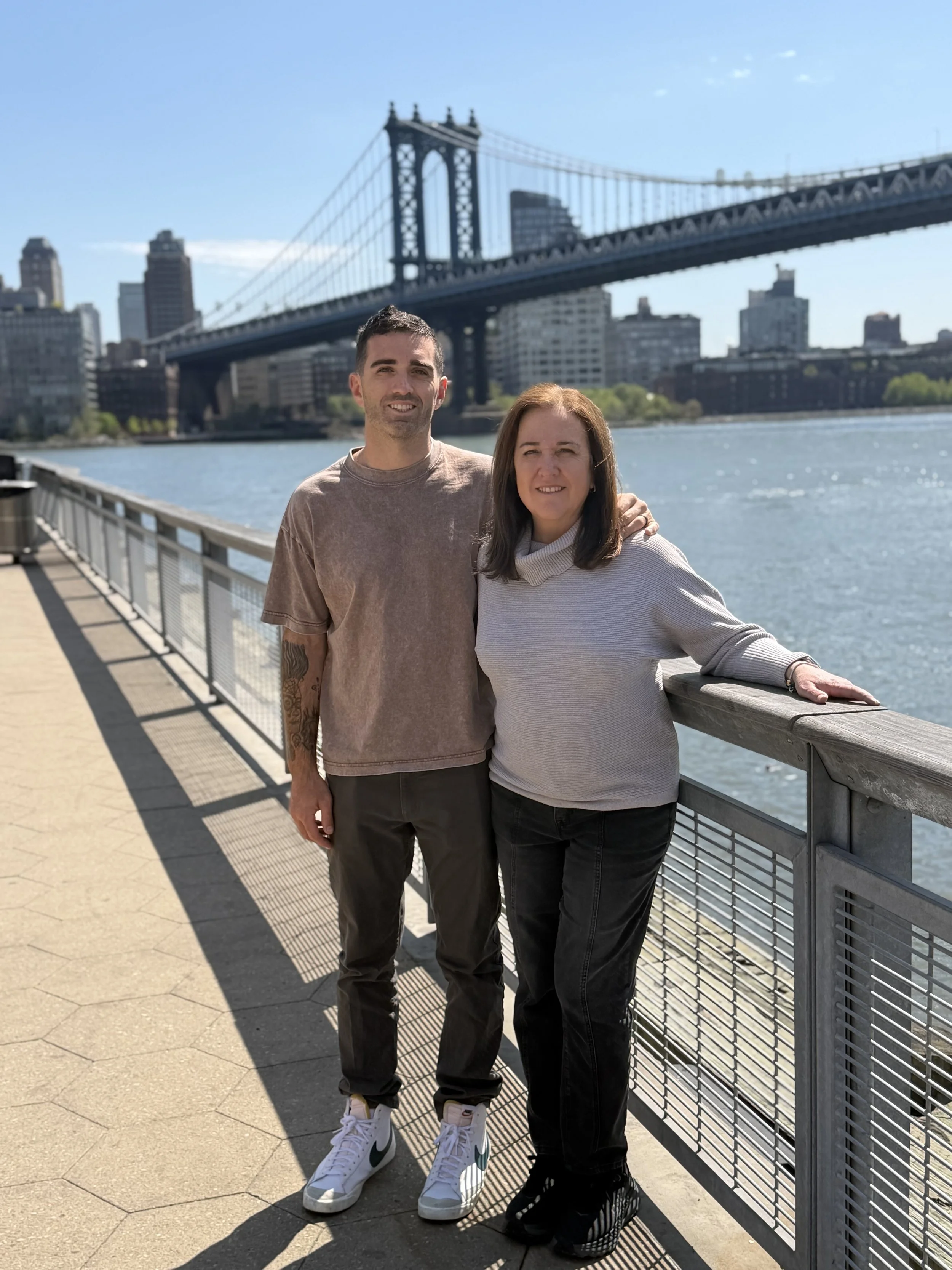 A man and a woman standing side by side on a waterfront promenade with a city skyline and a suspension bridge in the background. The man is wearing a tan t-shirt, black pants, and white sneakers, while the woman is wearing a light gray sweater, black pants, and black shoes. The man has his arm around the woman, and they are smiling at the camera on a sunny day.