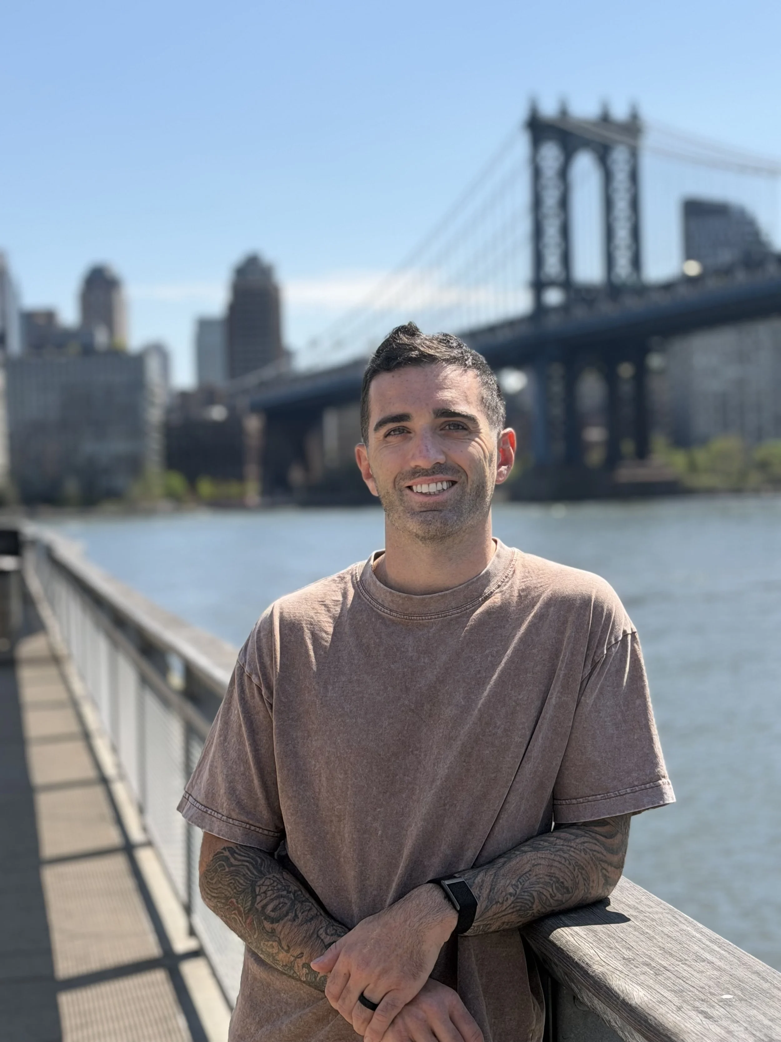 A man with short dark hair and tattoos on his arms, wearing a brown T-shirt, smiling while leaning on a wooden railing on a bridge, with the Brooklyn Bridge and city skyline in the background.