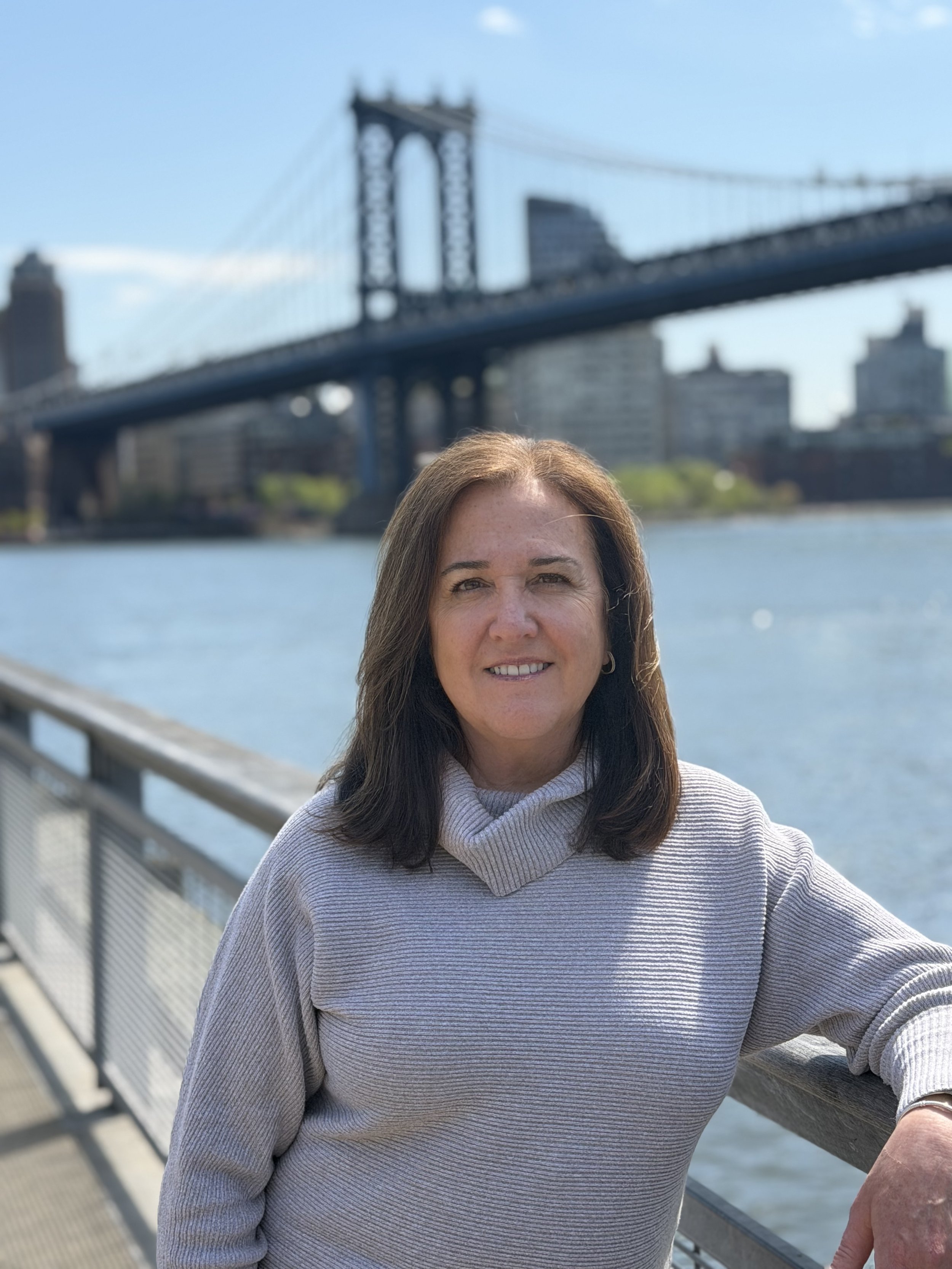 A woman with long brown hair smiling, wearing a light gray ribbed sweater, standing near a railing by water with a city skyline and the Manhattan Bridge in the background.