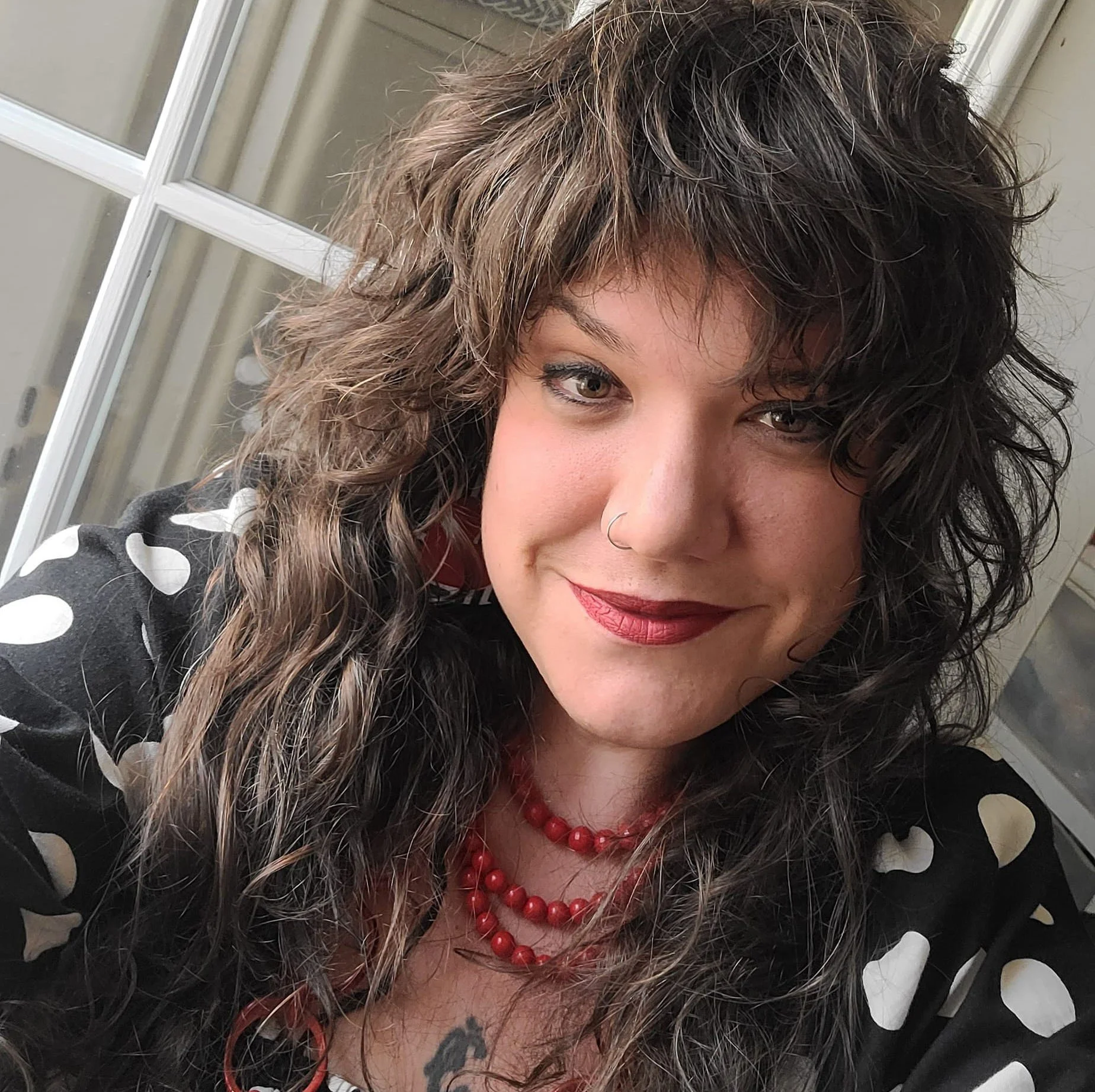 A woman with curly brown hair, wearing red lipstick, a nose ring, a black dress with white polka dots, and red jewelry, smiling in front of a window.