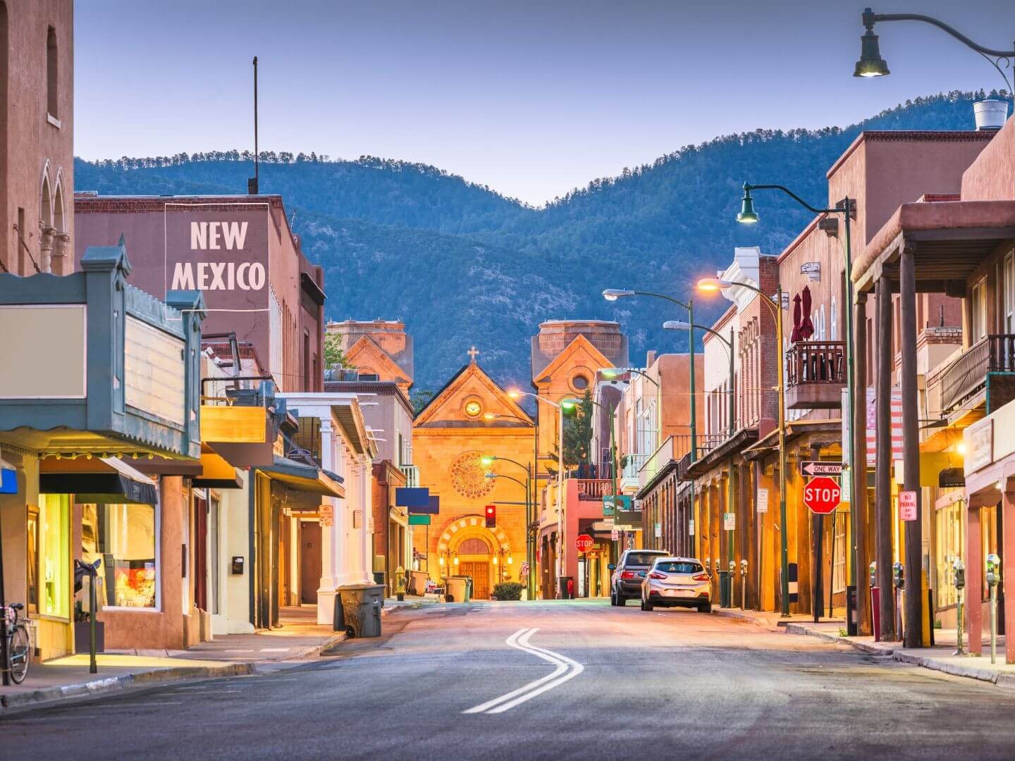 A quiet street in a small town with colorful buildings, street lamps, parked cars, and a church at the end of the street with mountains in the background.