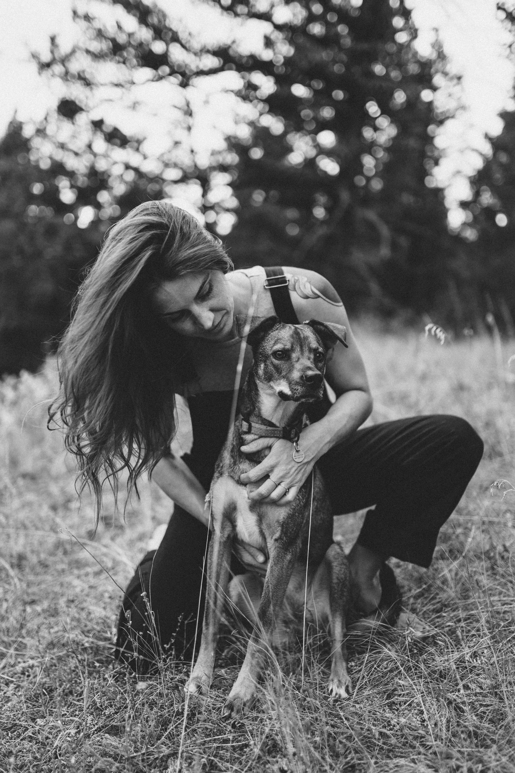 A woman with long hair and a tattoo on her shoulder, kneeling in a field and hugging a lean dog with a collar, in a black and white photograph.