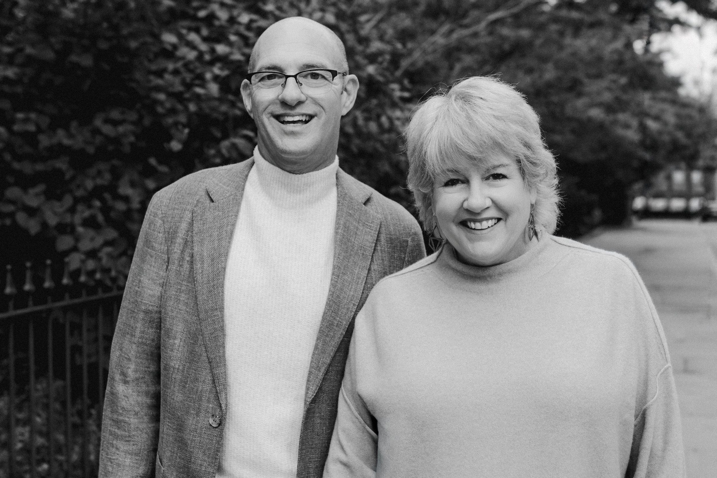 A black and white photo of a man and woman smiling outdoors, standing in front of bushes and a fence.