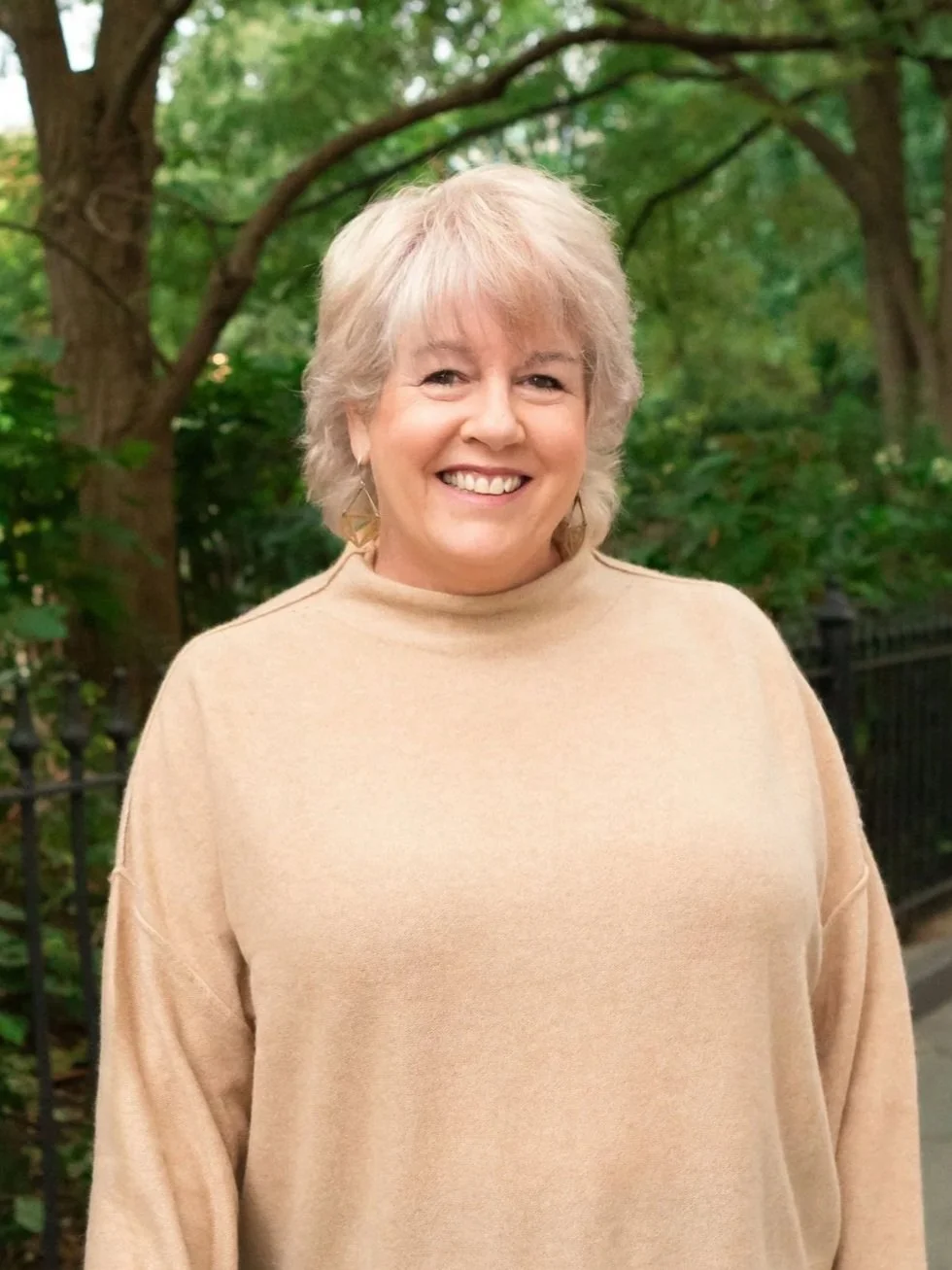A smiling woman with blonde hair, wearing a beige sweater, standing outdoors with green trees and a black metal fence in the background.