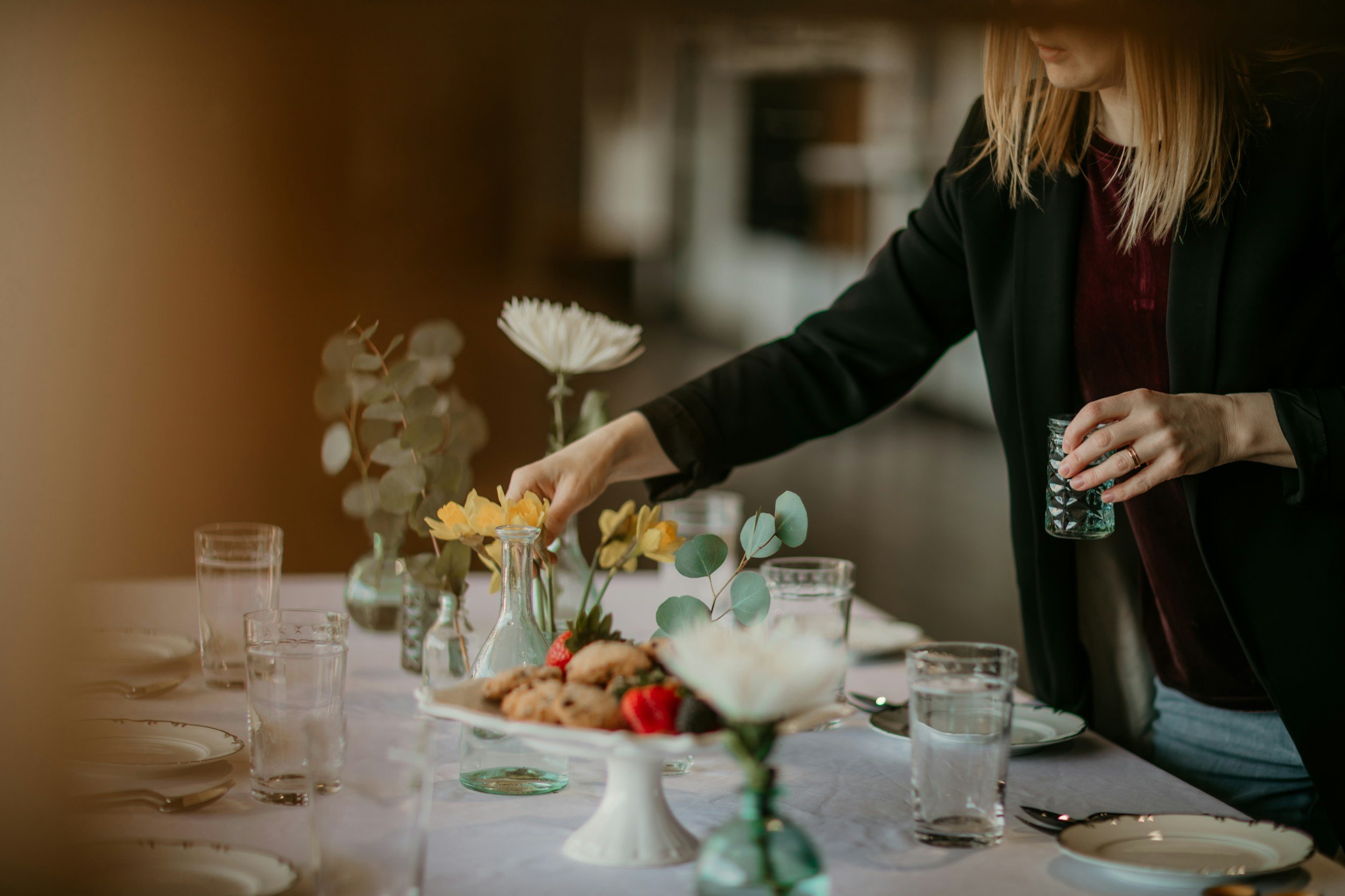 A woman arranging a table with a plate of cookies and strawberries, decorative vases with flowers and leaves, and glasses of water.