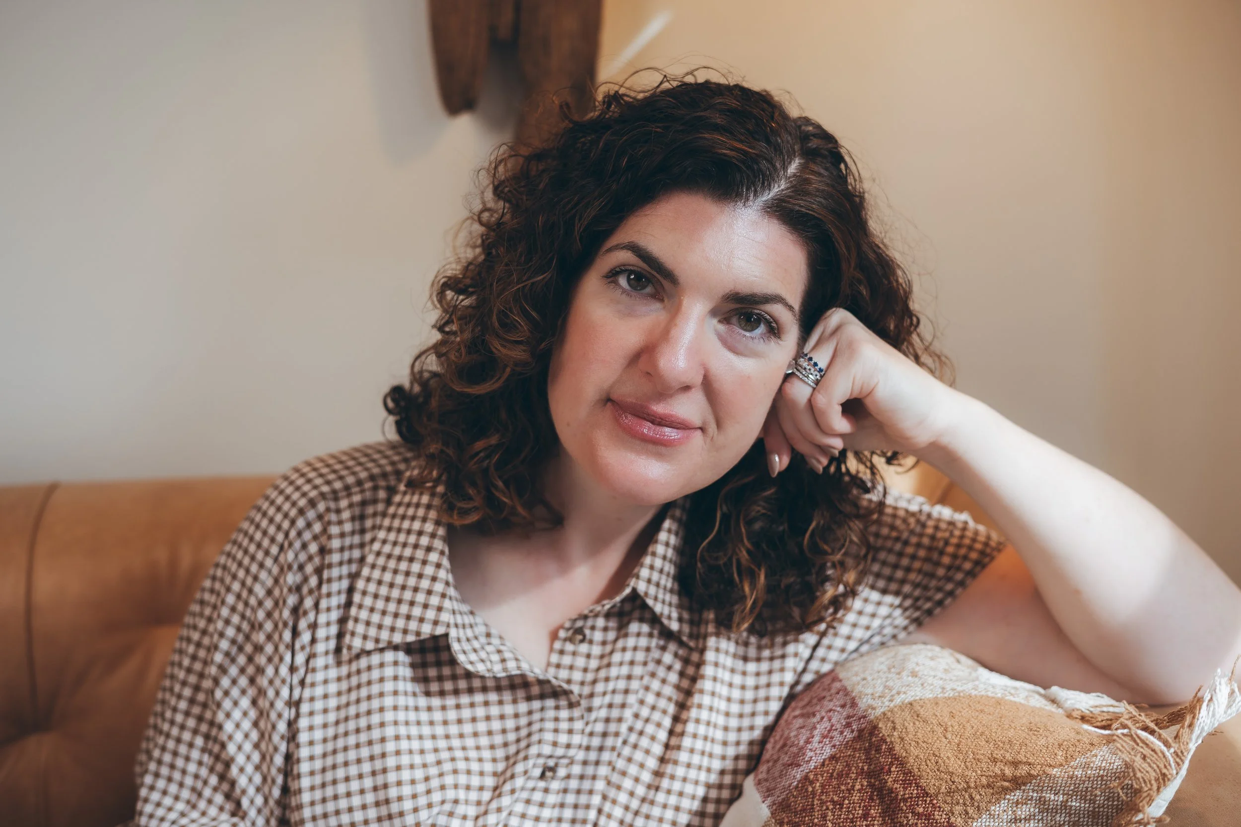 A woman with curly brown hair, wearing a checkered shirt, sitting on a tan leather couch, resting her head on her hand, and looking at the camera.